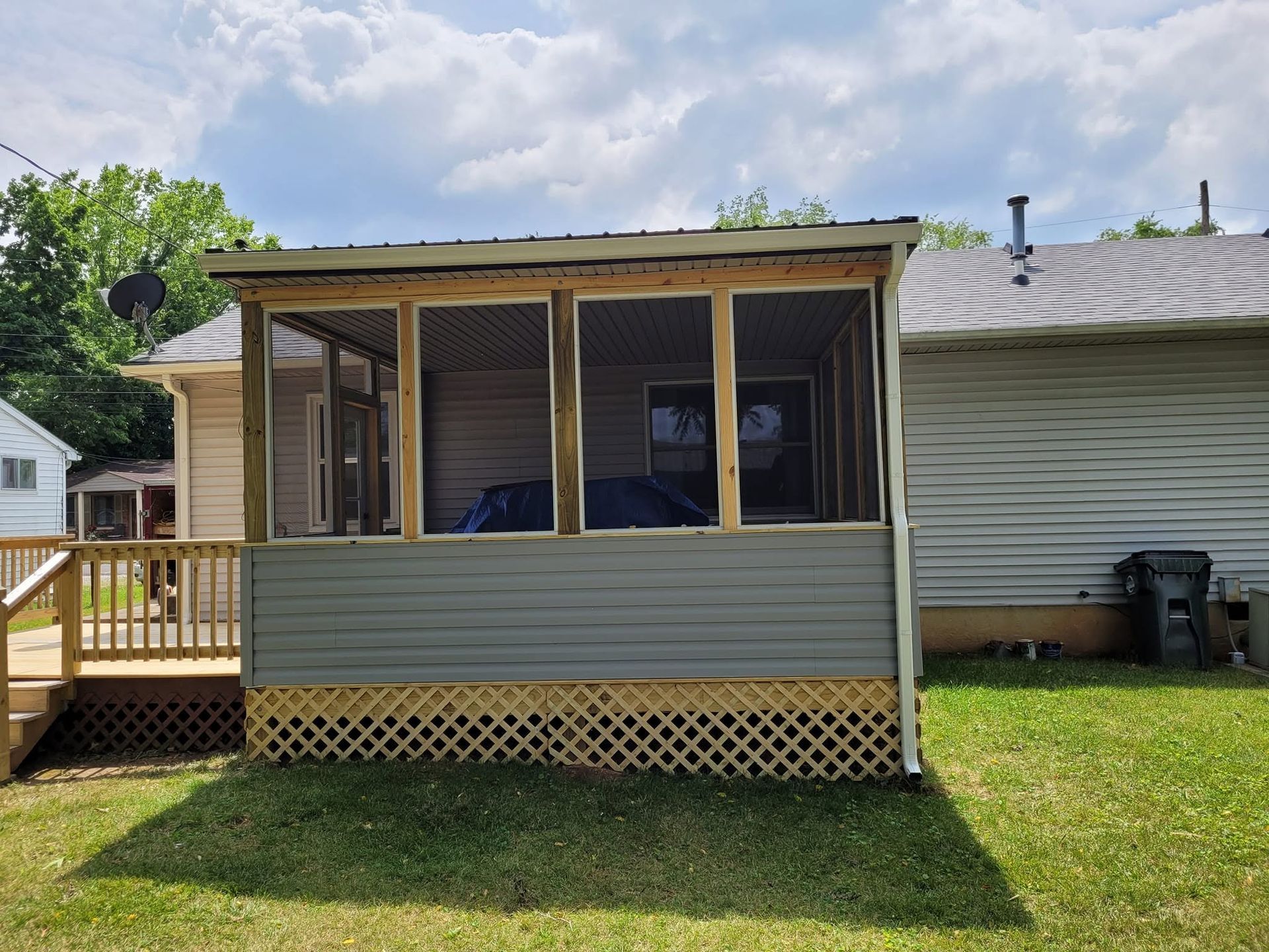 Screened porch with beige siding, wooden railing, and lattice skirt attached to a house on a grassy lawn