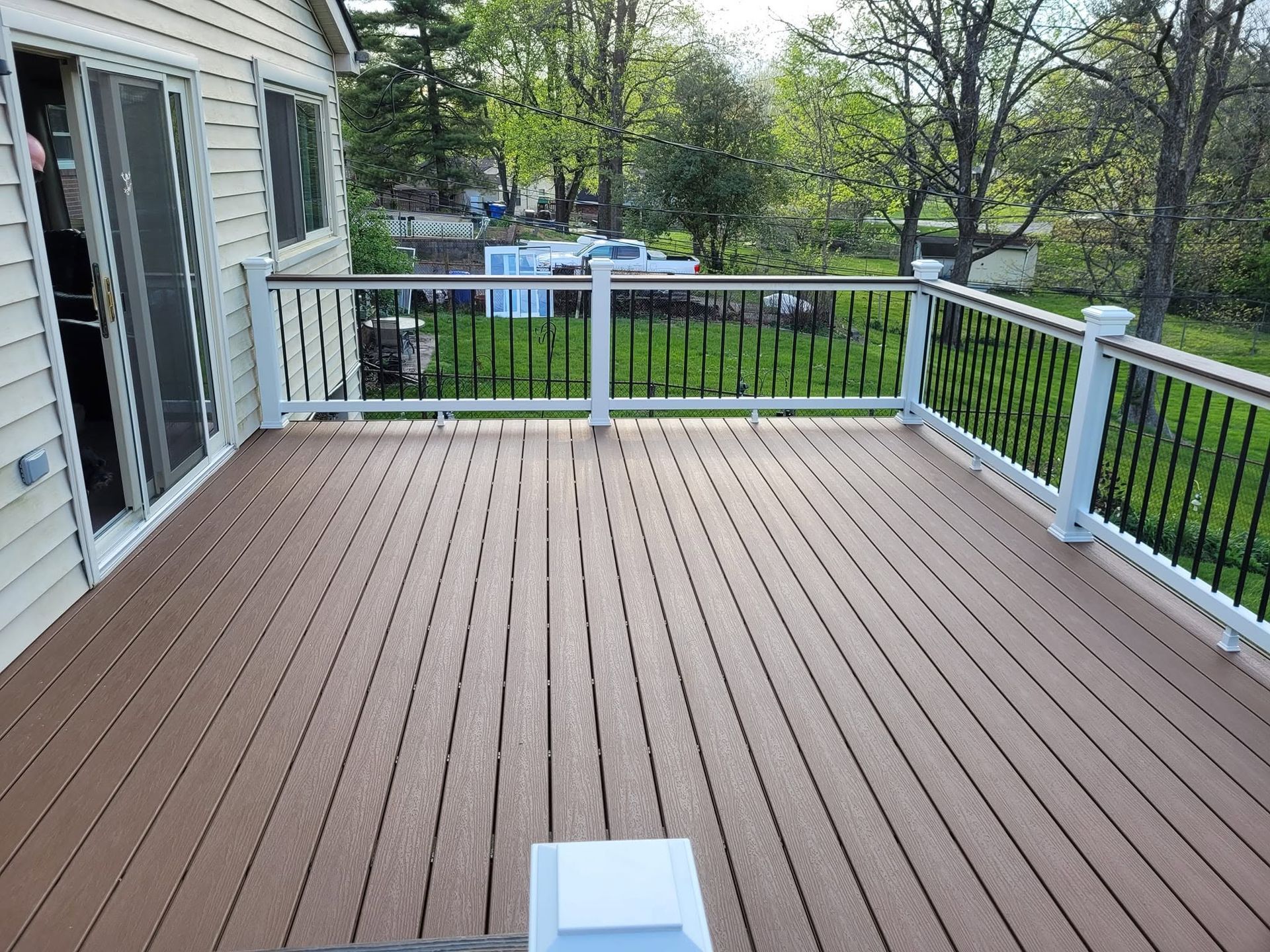 Empty wooden deck with white railings and a house door, overlooking a green yard and trees.