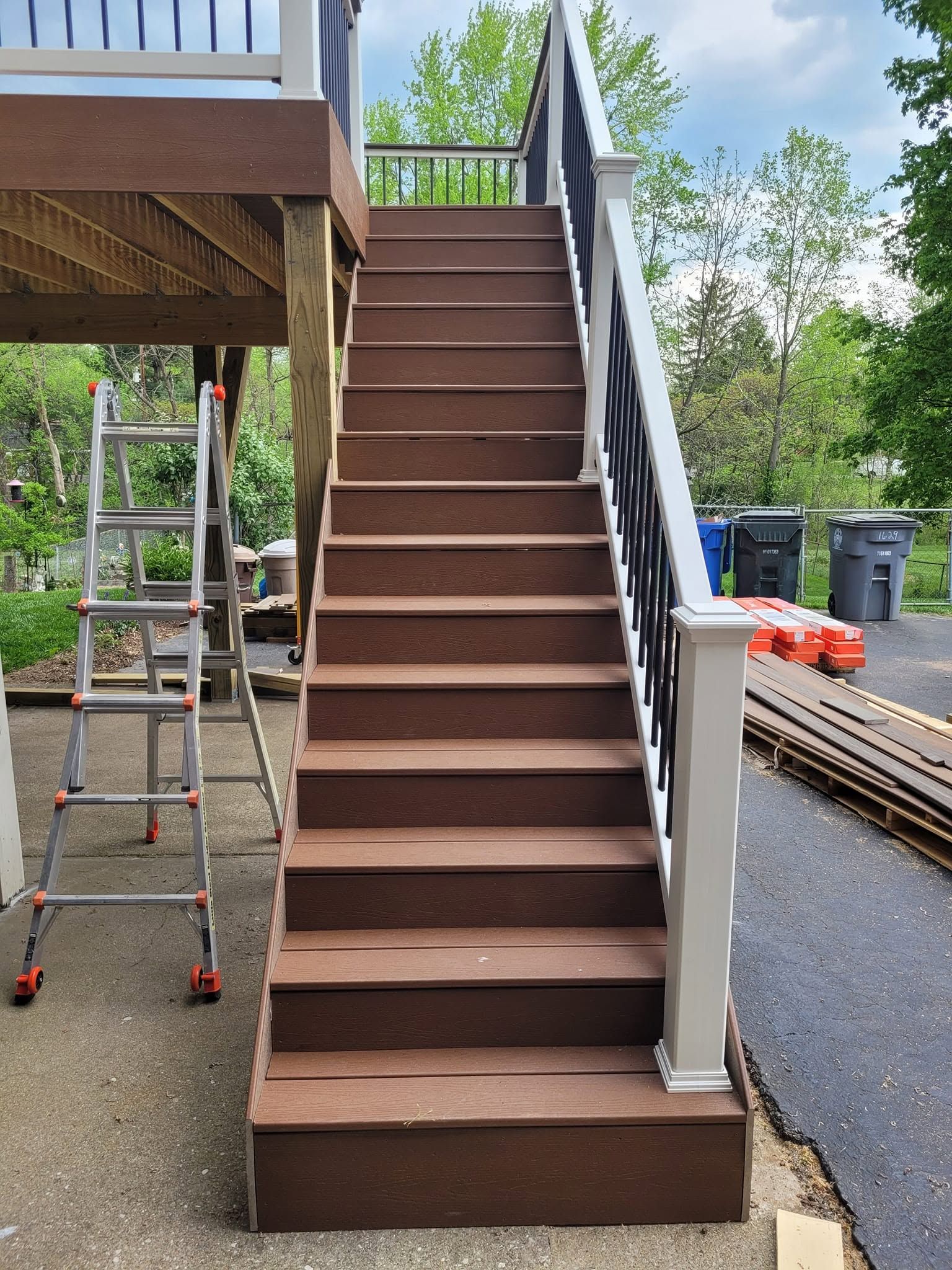 Brown outdoor staircase beside a deck, with a ladder on the left and gravel driveway on the right.