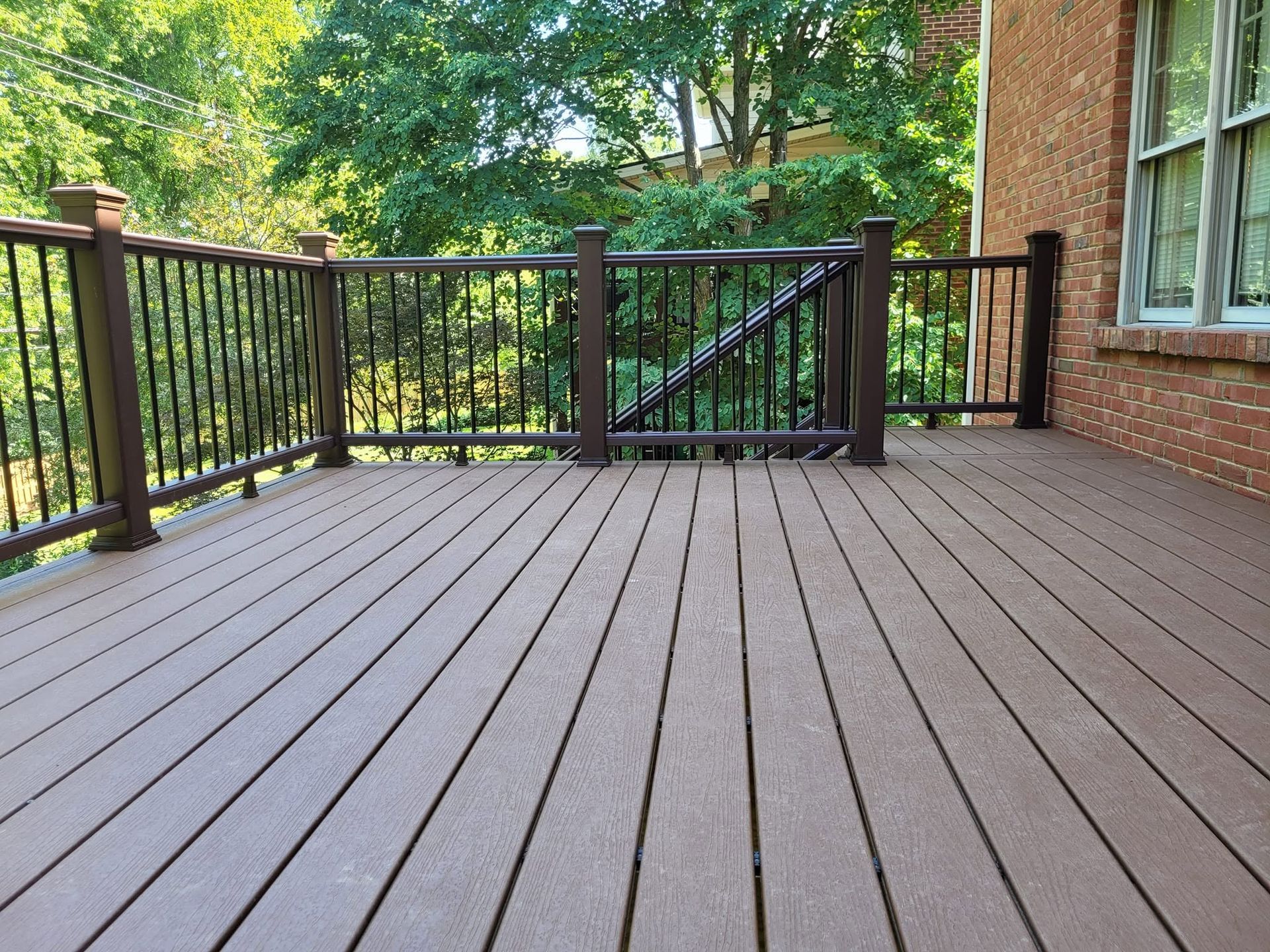 Wooden deck with black railings and a gate overlooking green trees beside a brick house