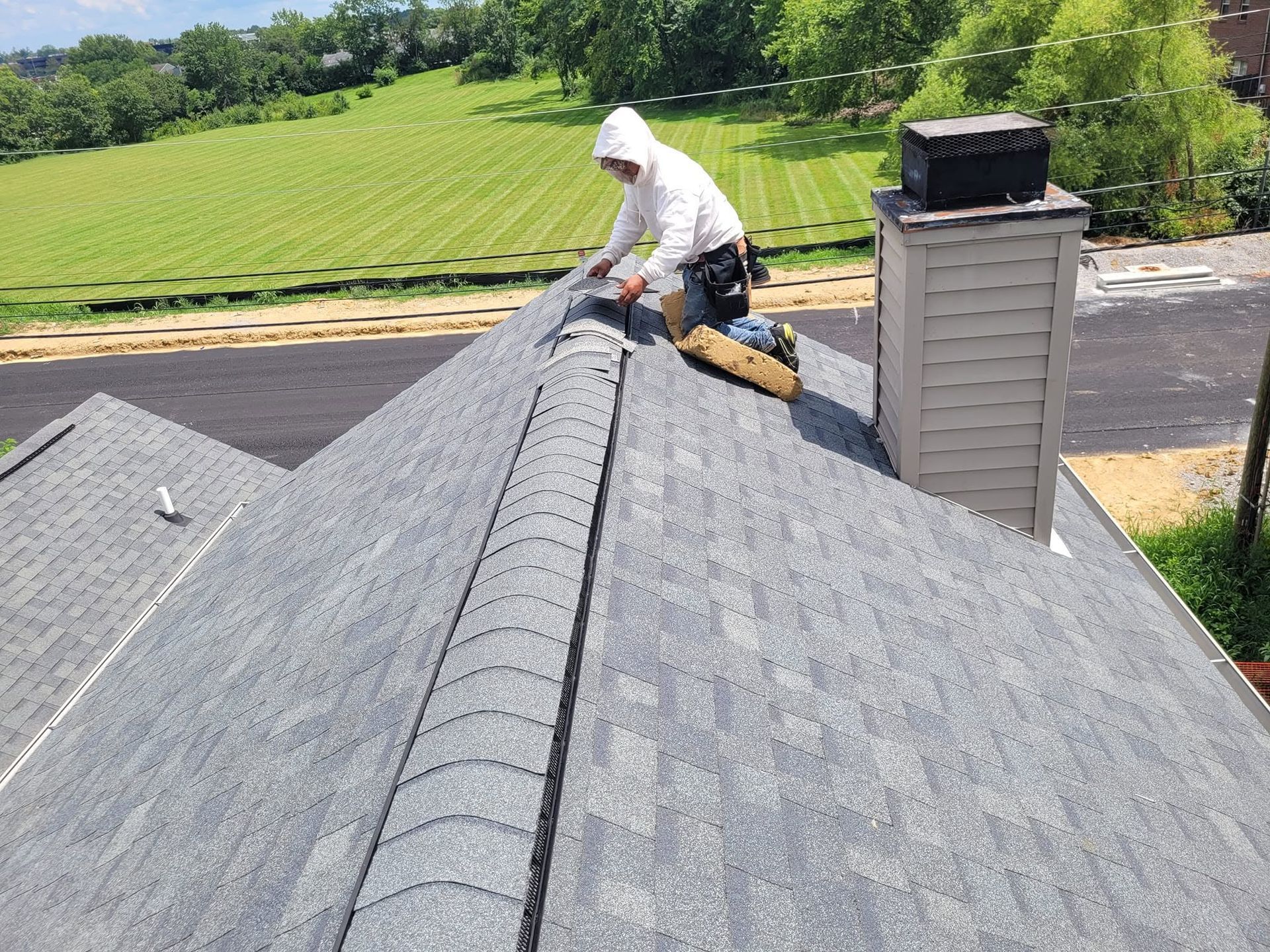 Worker installing shingles on a gray roof beside a chimney on a sunny day