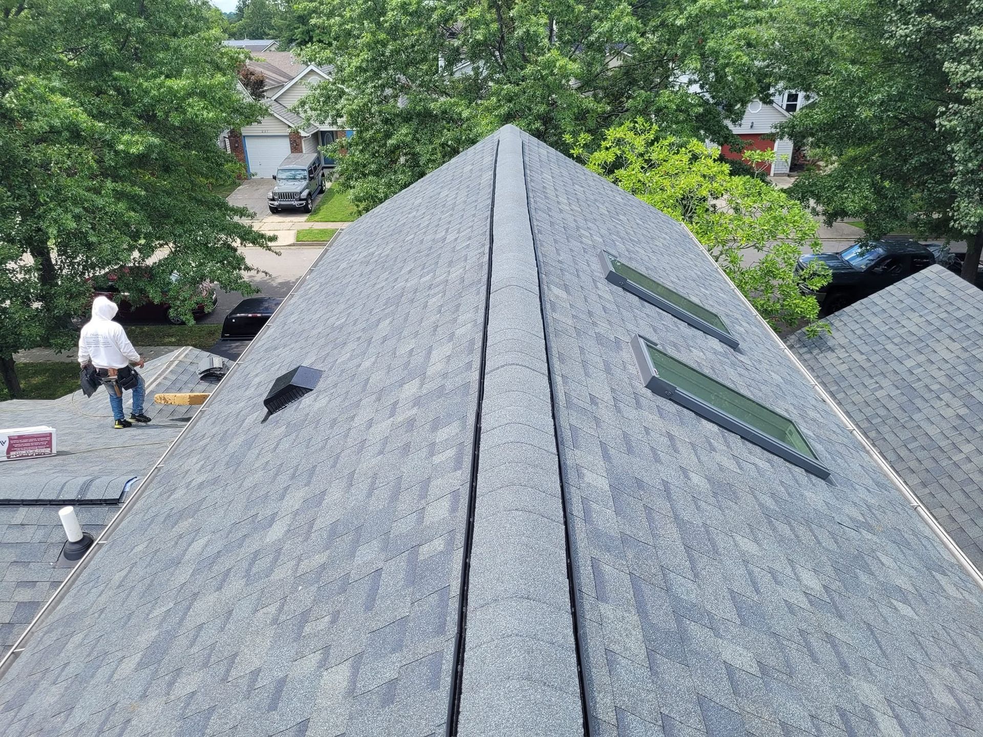 Steep gray-shingled roof with skylights, viewed from above, with trees and a person in the background
