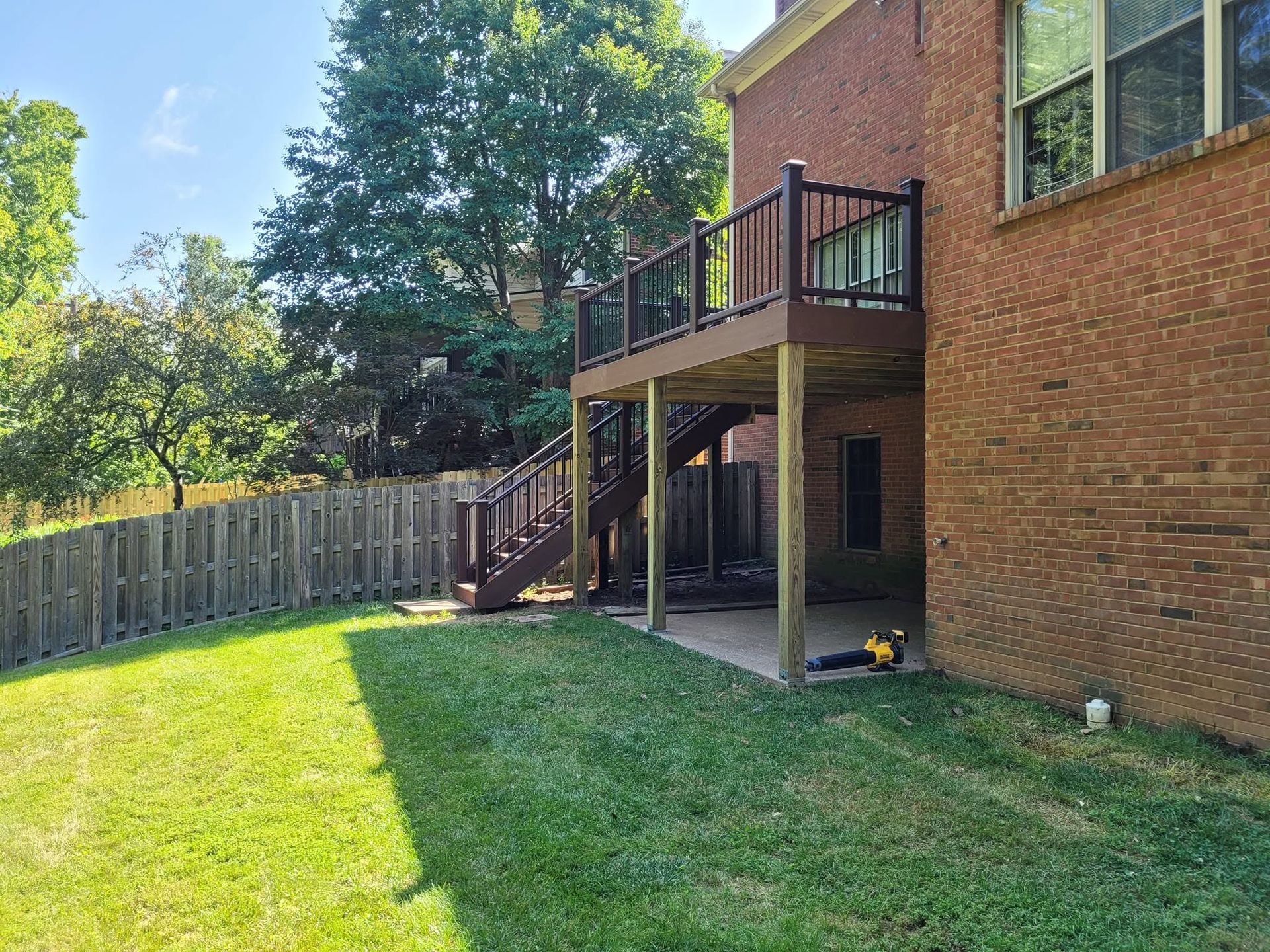 Backyard with green lawn, wooden deck stairs, brick house, and fenced yard in bright sunlight