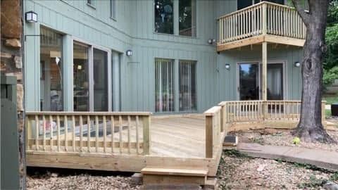 Wooden apartment deck and ramp with railings beside a beige building and large tree outdoors