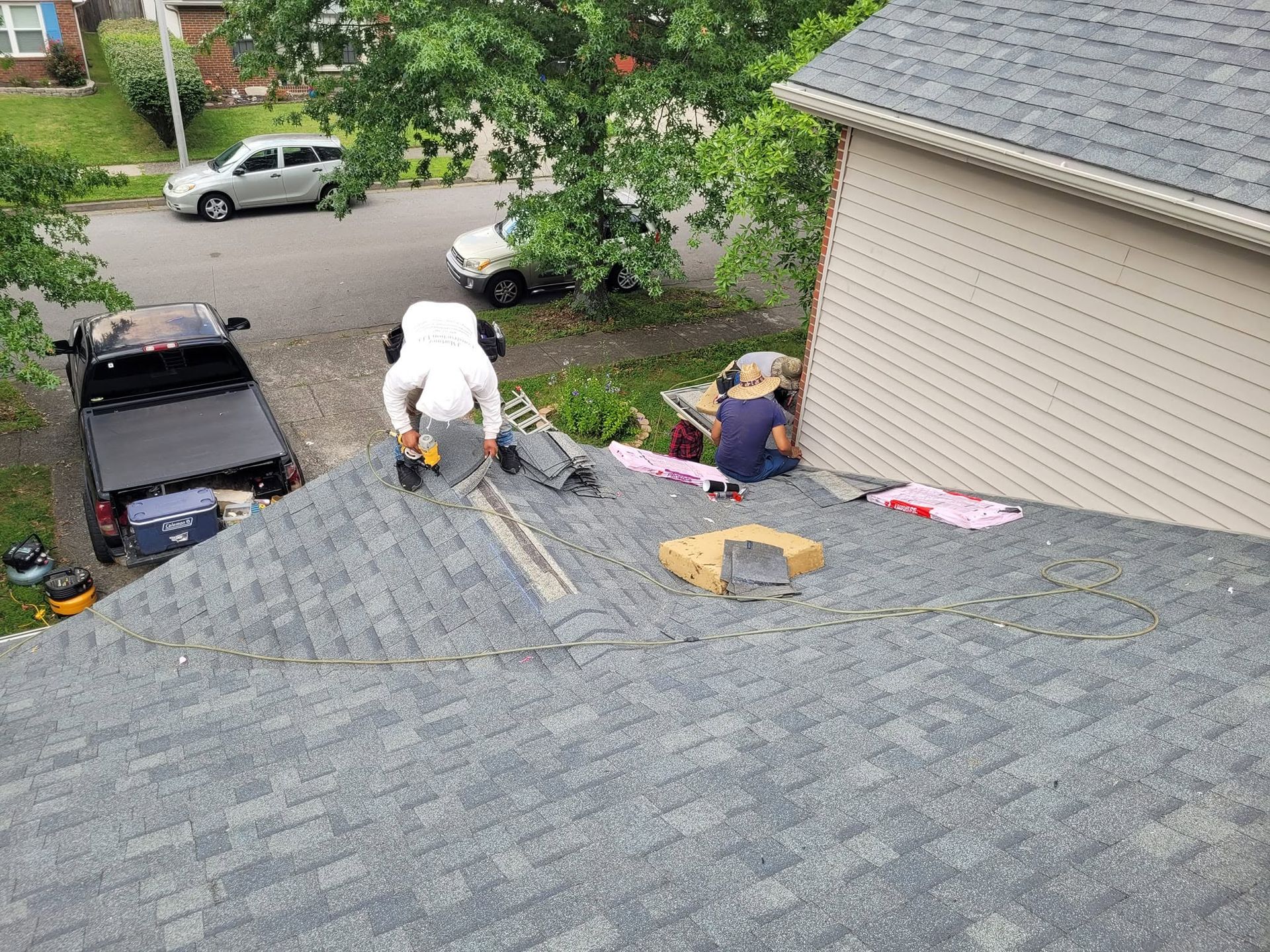 Roofers working on a gray shingled roof beside a beige house, with tools and materials spread out.