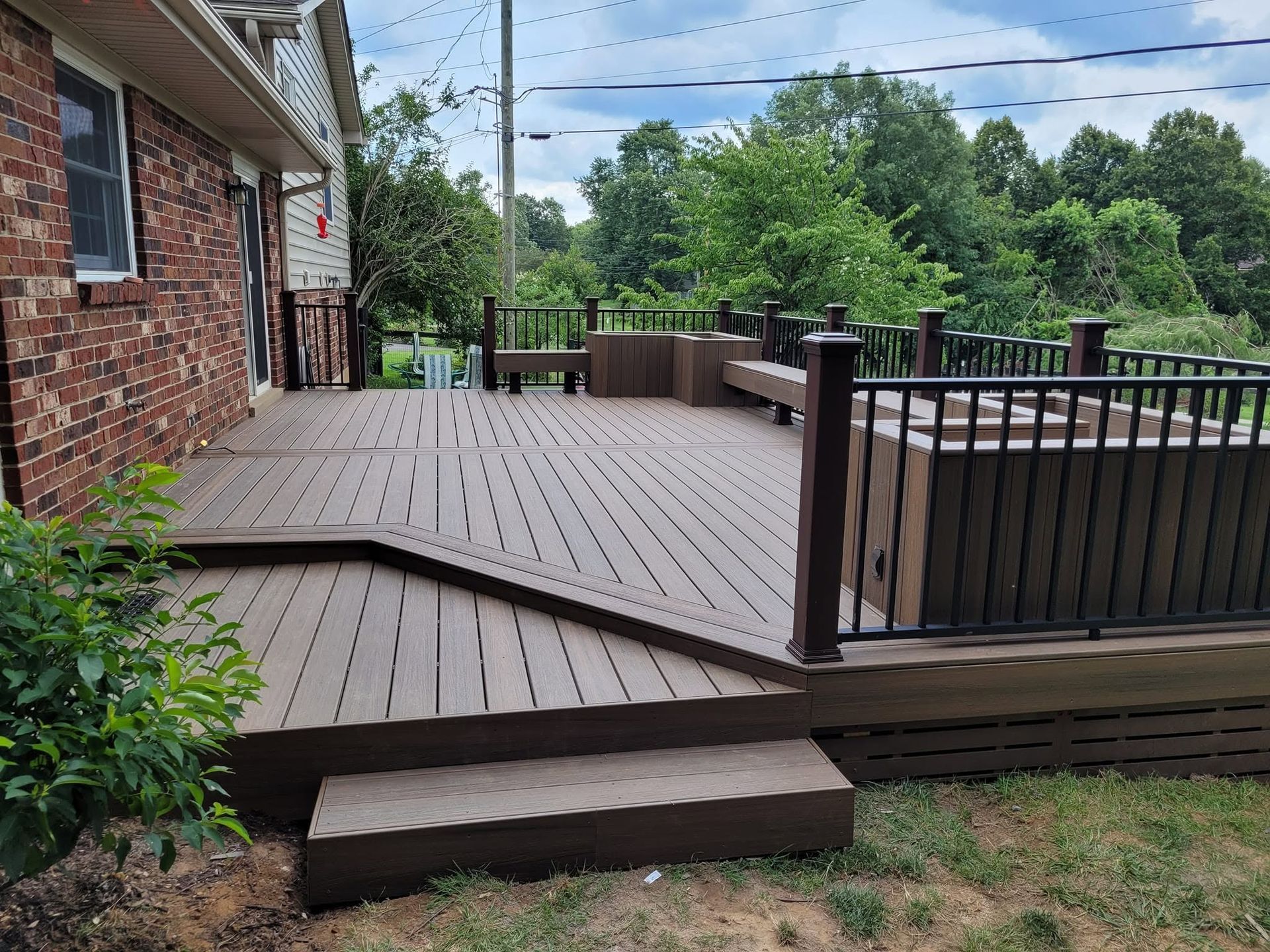 Raised wooden deck beside brick house, with steps, railing, and green trees beyond.
