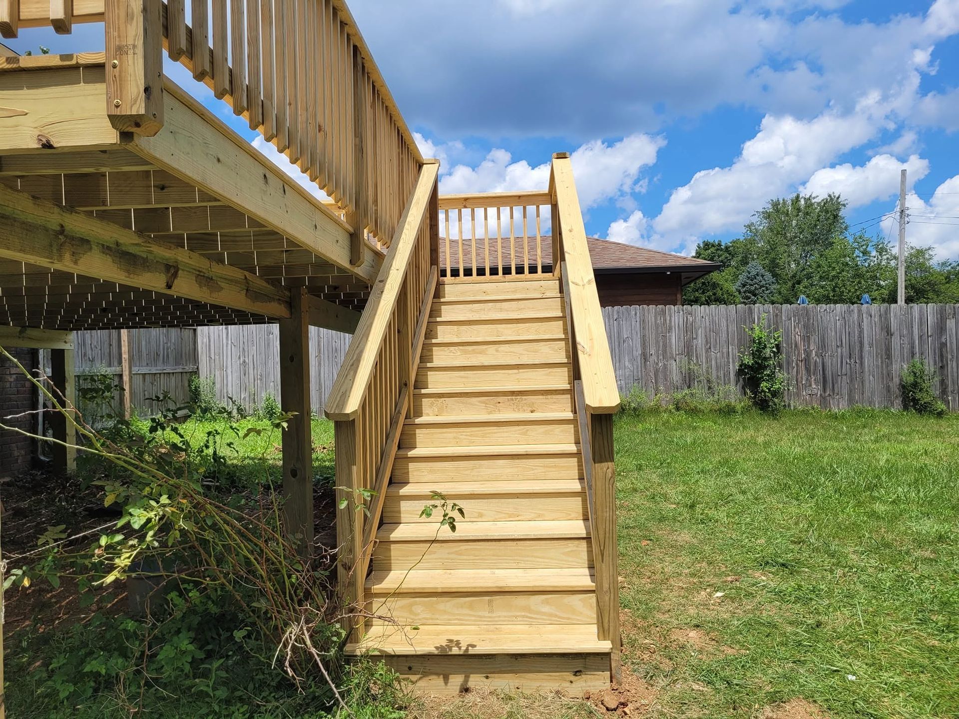 Wooden backyard staircase leading to an elevated deck beside a fenced lawn under a blue sky