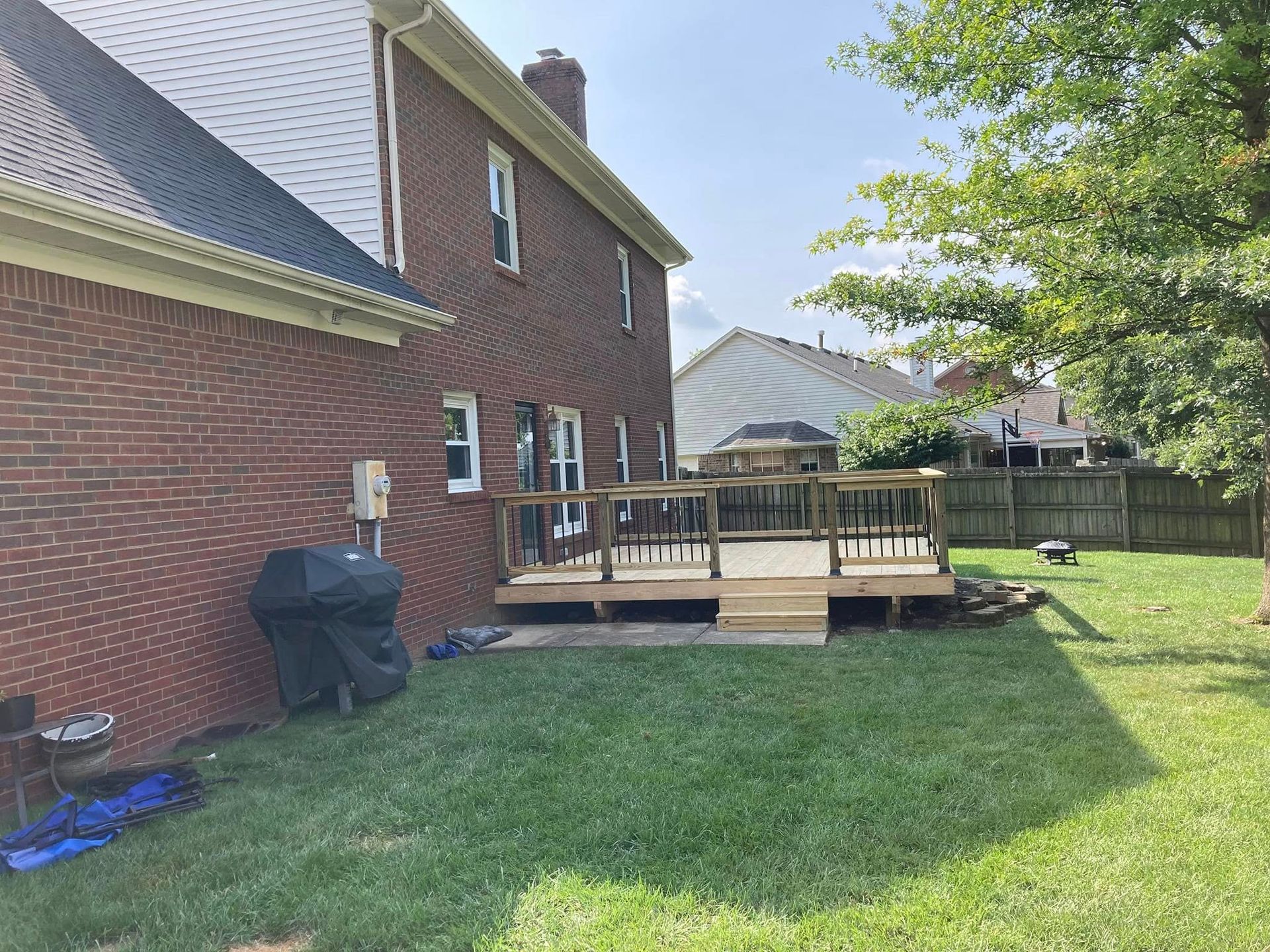 Backyard deck beside a red brick house, with green lawn, fence, and trees in the background.