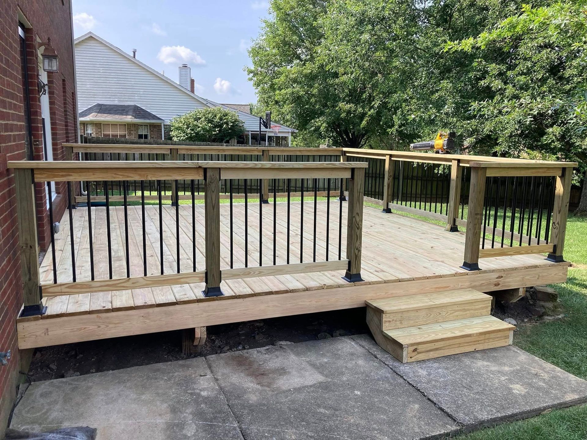 Wooden backyard deck with railing and steps, set beside a house and driveway under a leafy tree.