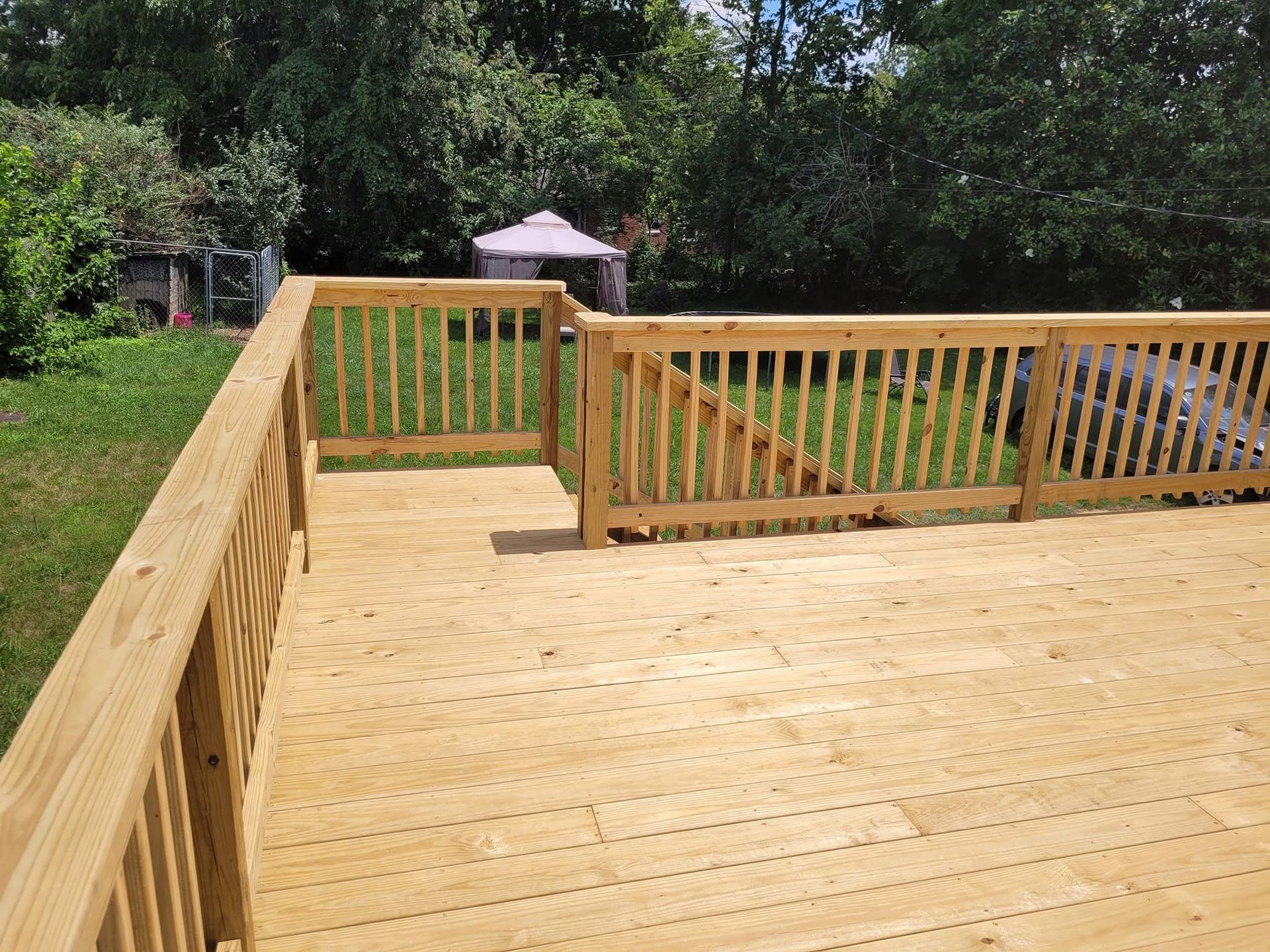 Wooden deck with railings in a sunny backyard, bordered by trees and a patio umbrella in the distance
