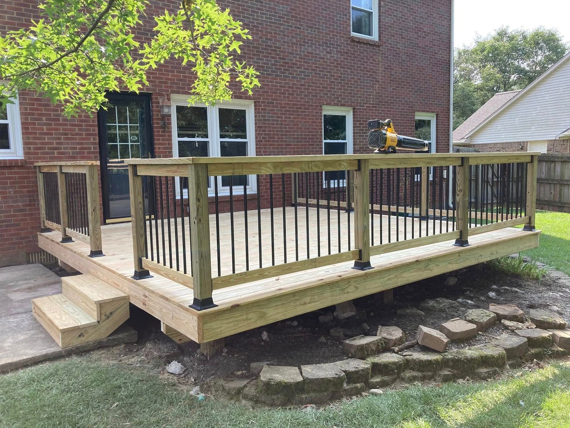Wooden backyard deck with railing attached to a brick house, with steps and a stone patio below