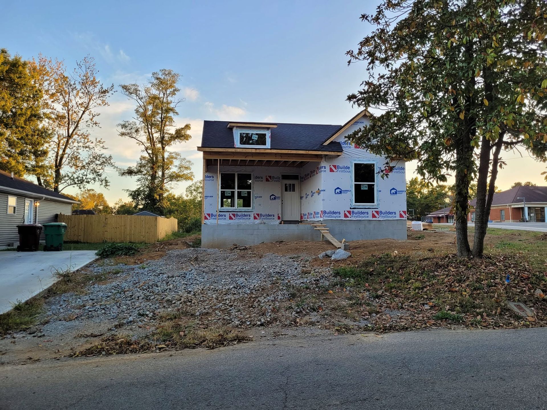 Small house under construction with gravel driveway, autumn trees, and a roadside in front