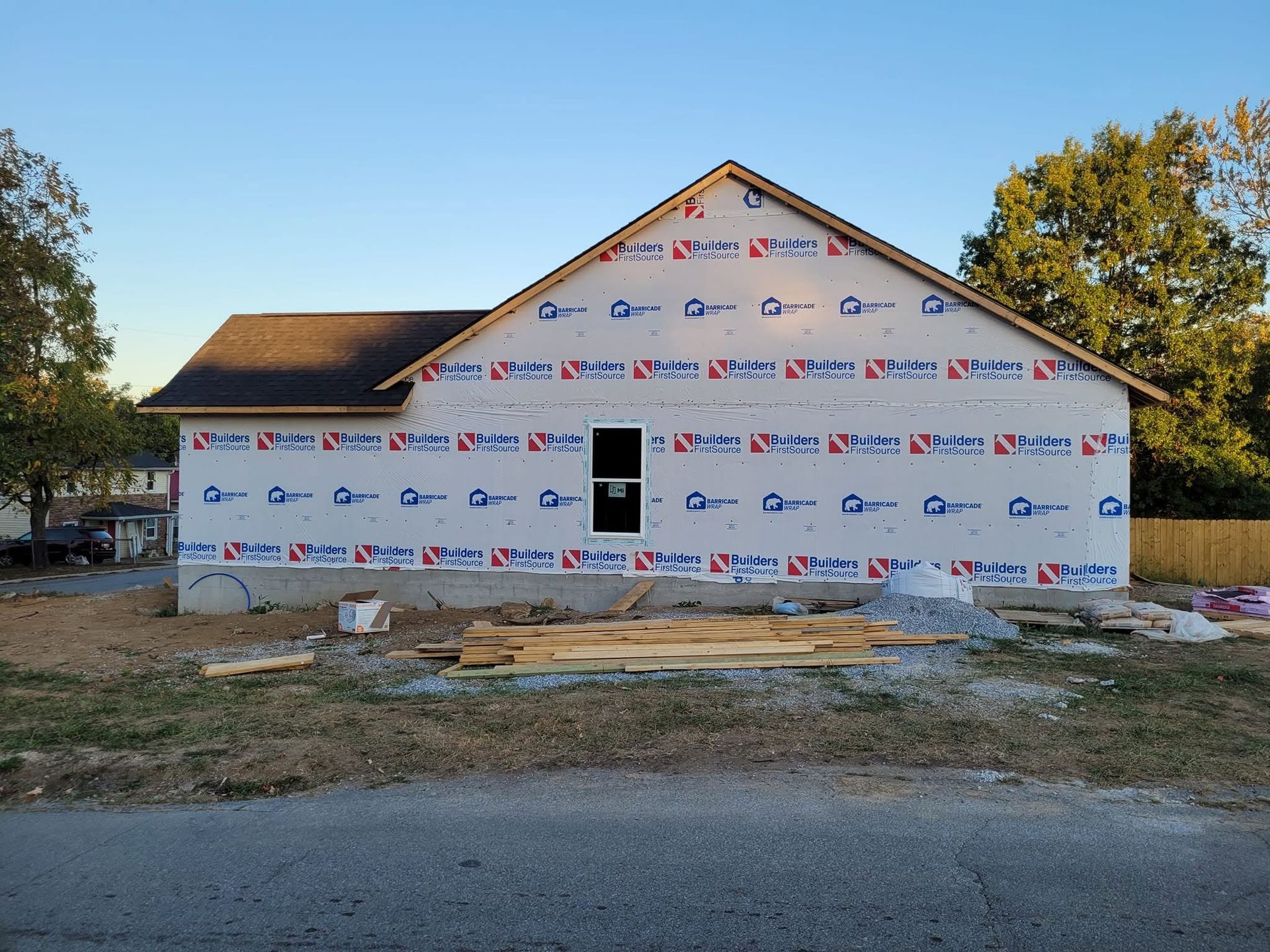 House under construction wrapped in white house wrap, with a front door opening and unfinished yard.