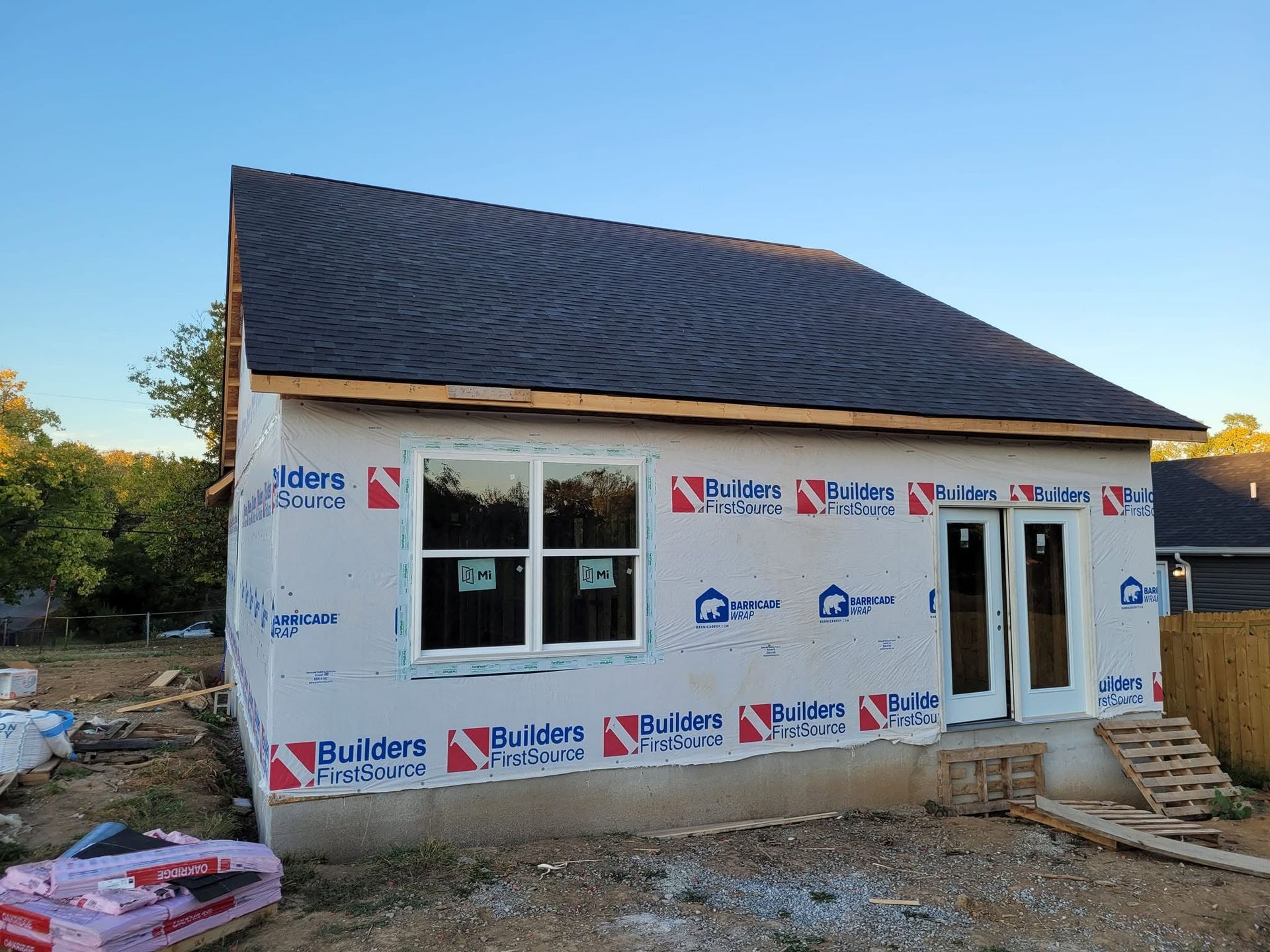 Small house under construction with roof shingles, wrapped in building material, at a dirt lot.