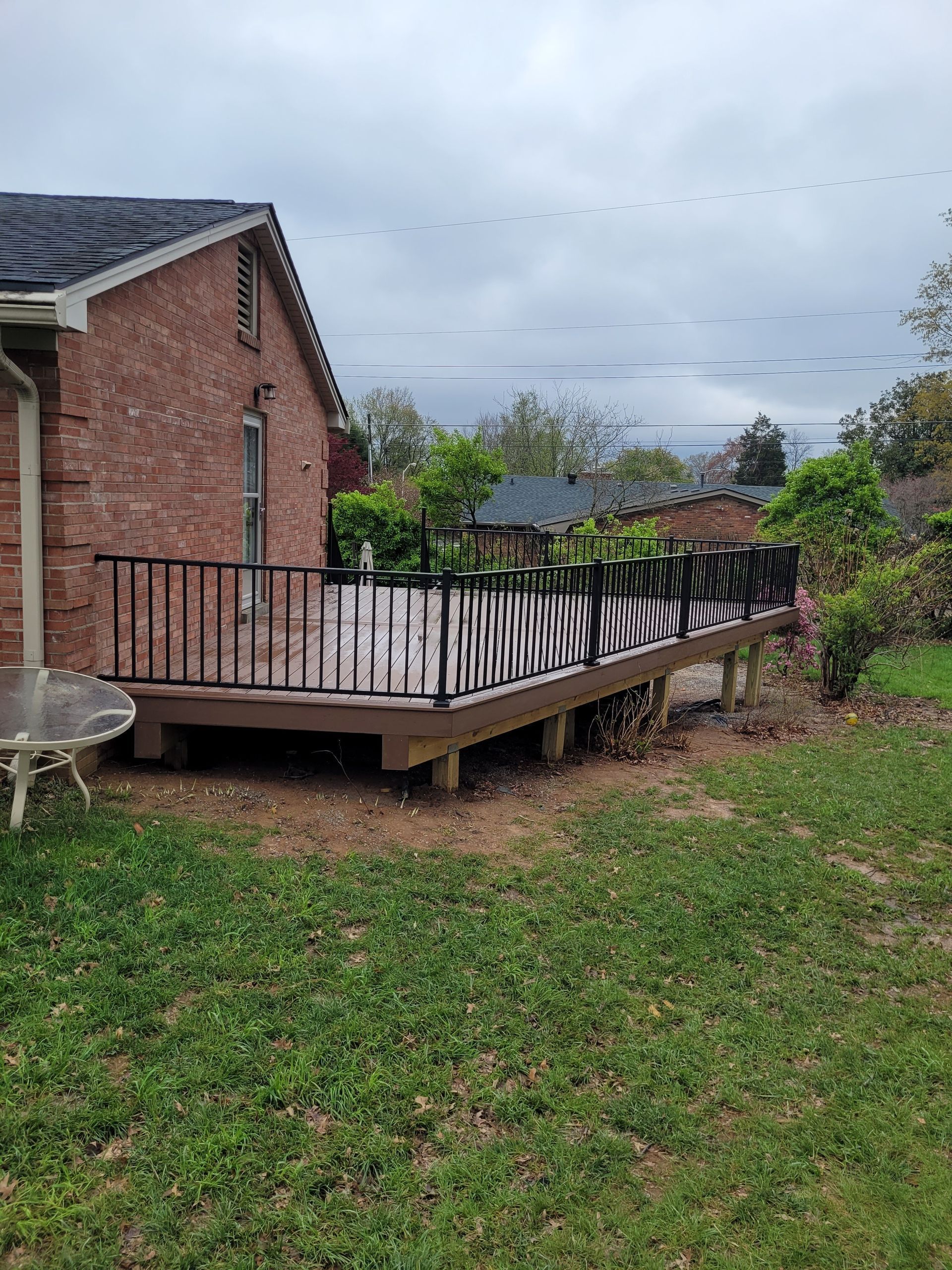 Backyard deck beside a brick house, with bent railings and a grassy yard under cloudy skies