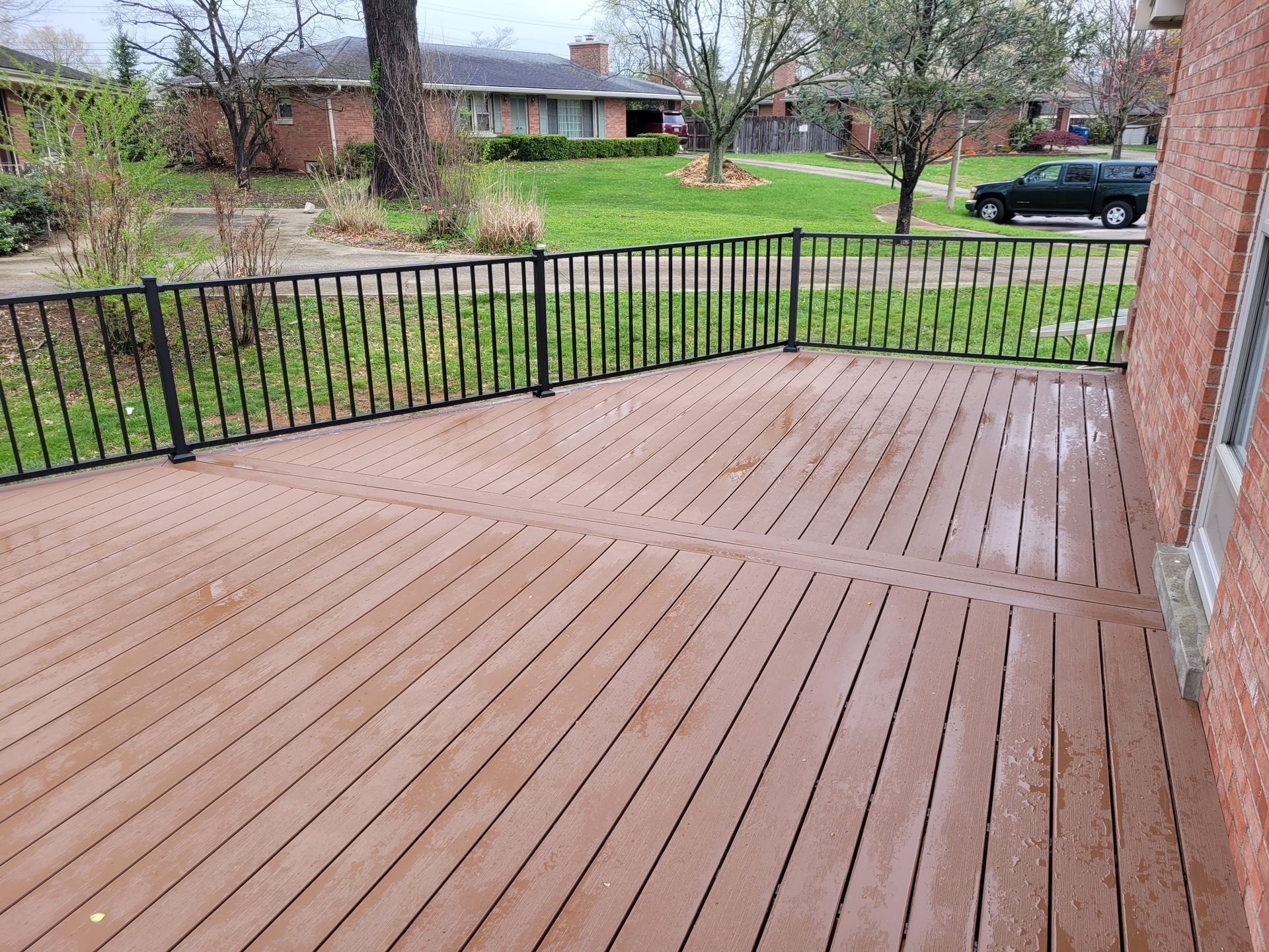 Wooden backyard deck with black railing beside a brick house, overlooking a grassy yard and parked car.