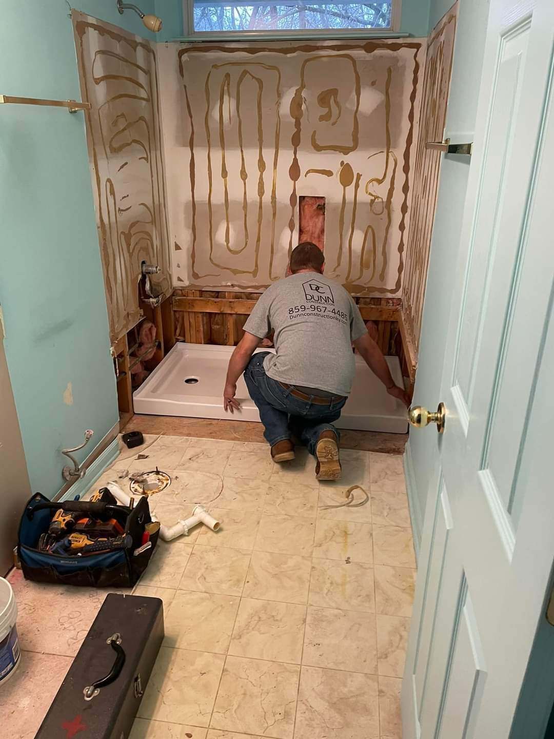 Person installing a bathtub surround in a small bathroom with exposed adhesive and tools on the floor