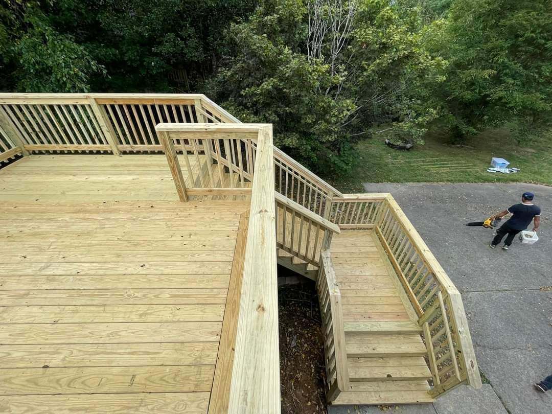Wooden deck with stairs and railing beside a gravel driveway, where a person is sitting in a wheelchair.