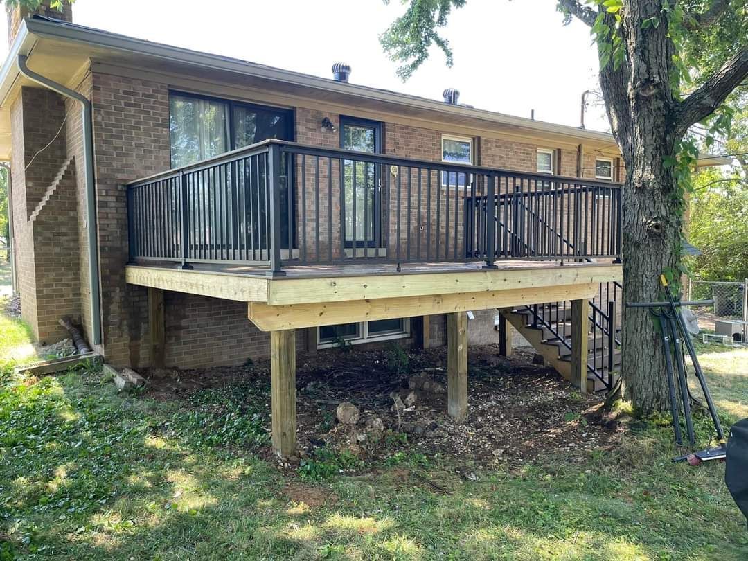 Raised backyard deck with black railing attached to a brick house, shaded by a large tree.