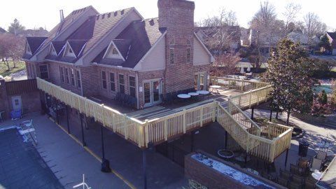 A large brick house with a wraparound porch and deck on a sunny day, viewed from above.