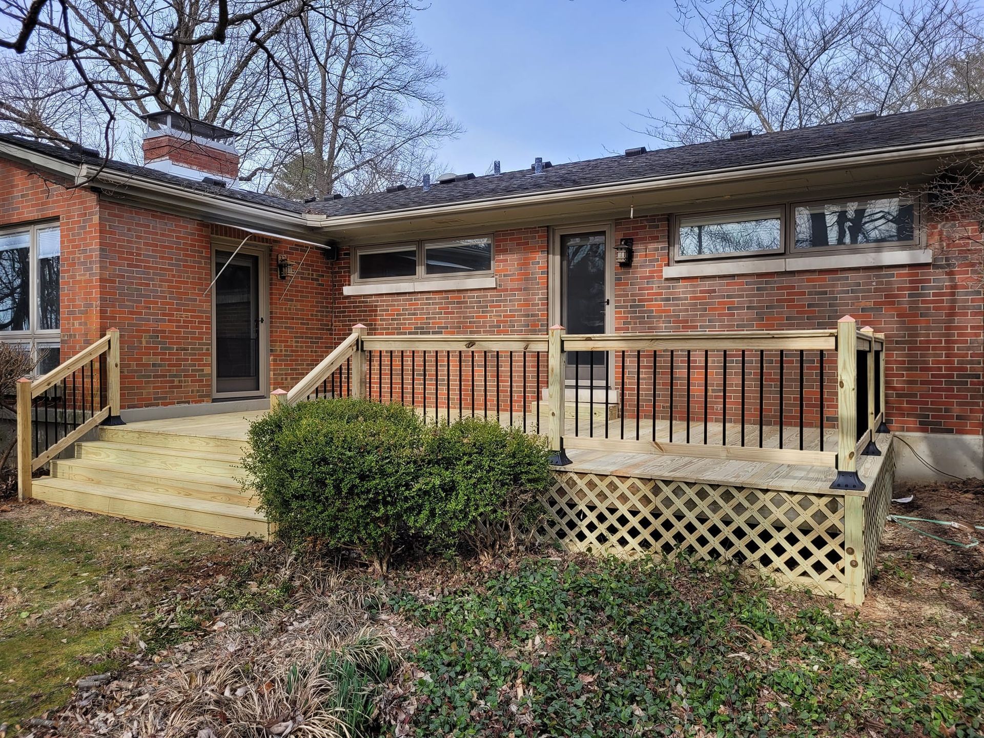 Brick house front entrance with wooden ramp, porch railing, and winter shrubs