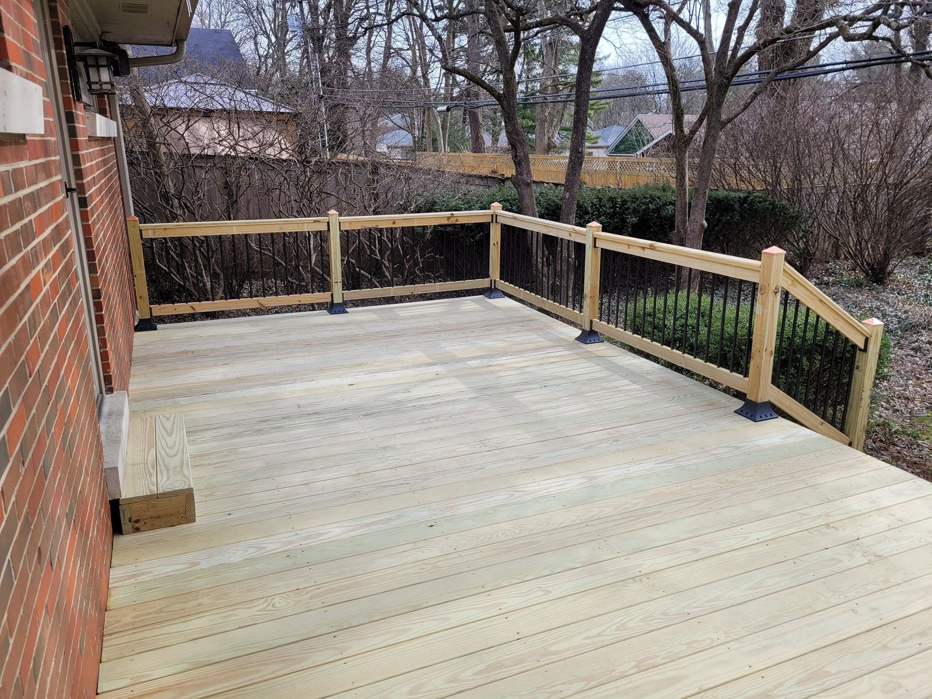 Wooden backyard deck with light railing beside a brick house and bare trees beyond.