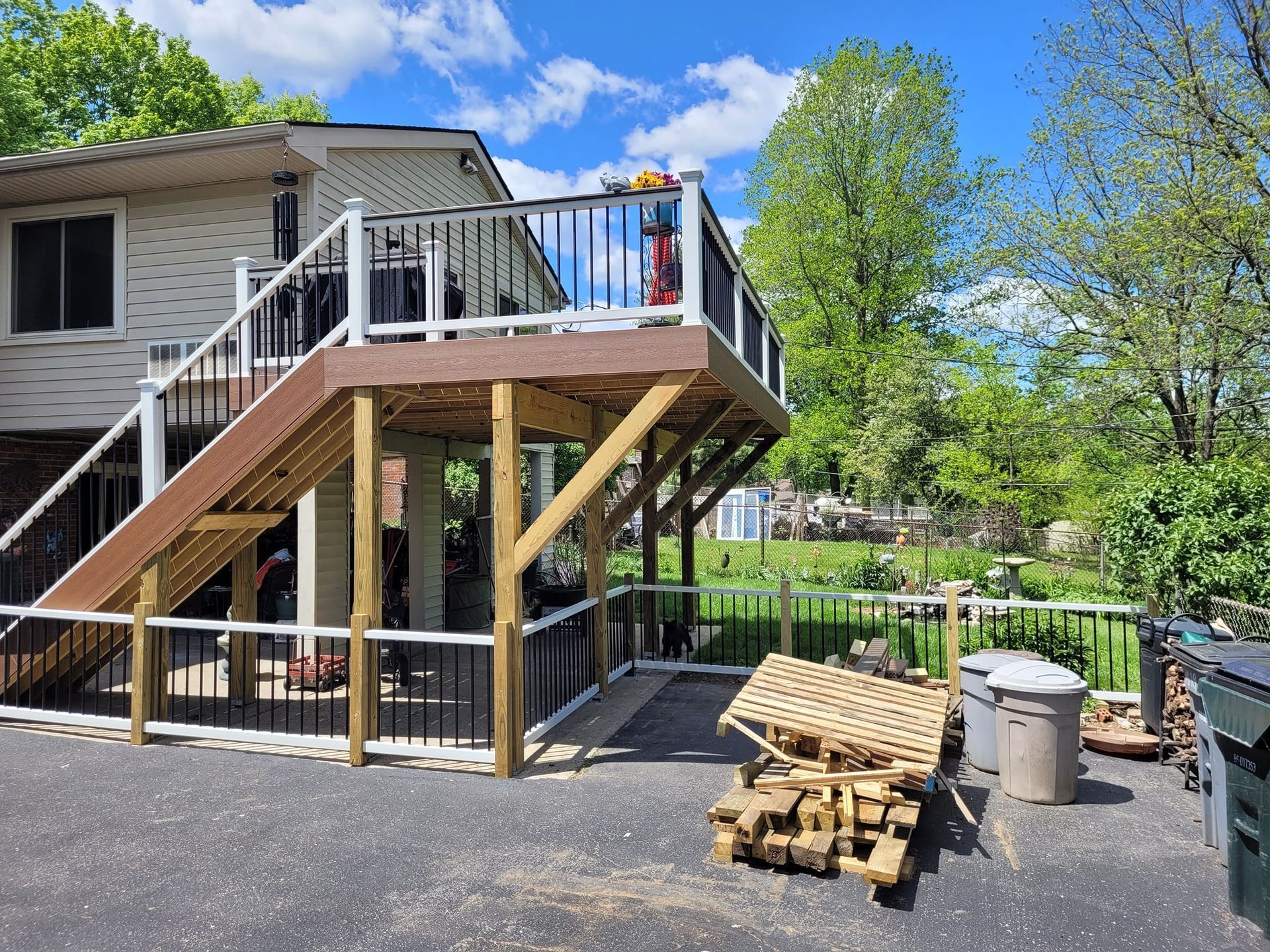 Raised deck and stairs on a house, with stacked firewood and trash bins in a gravel yard under a blue sky