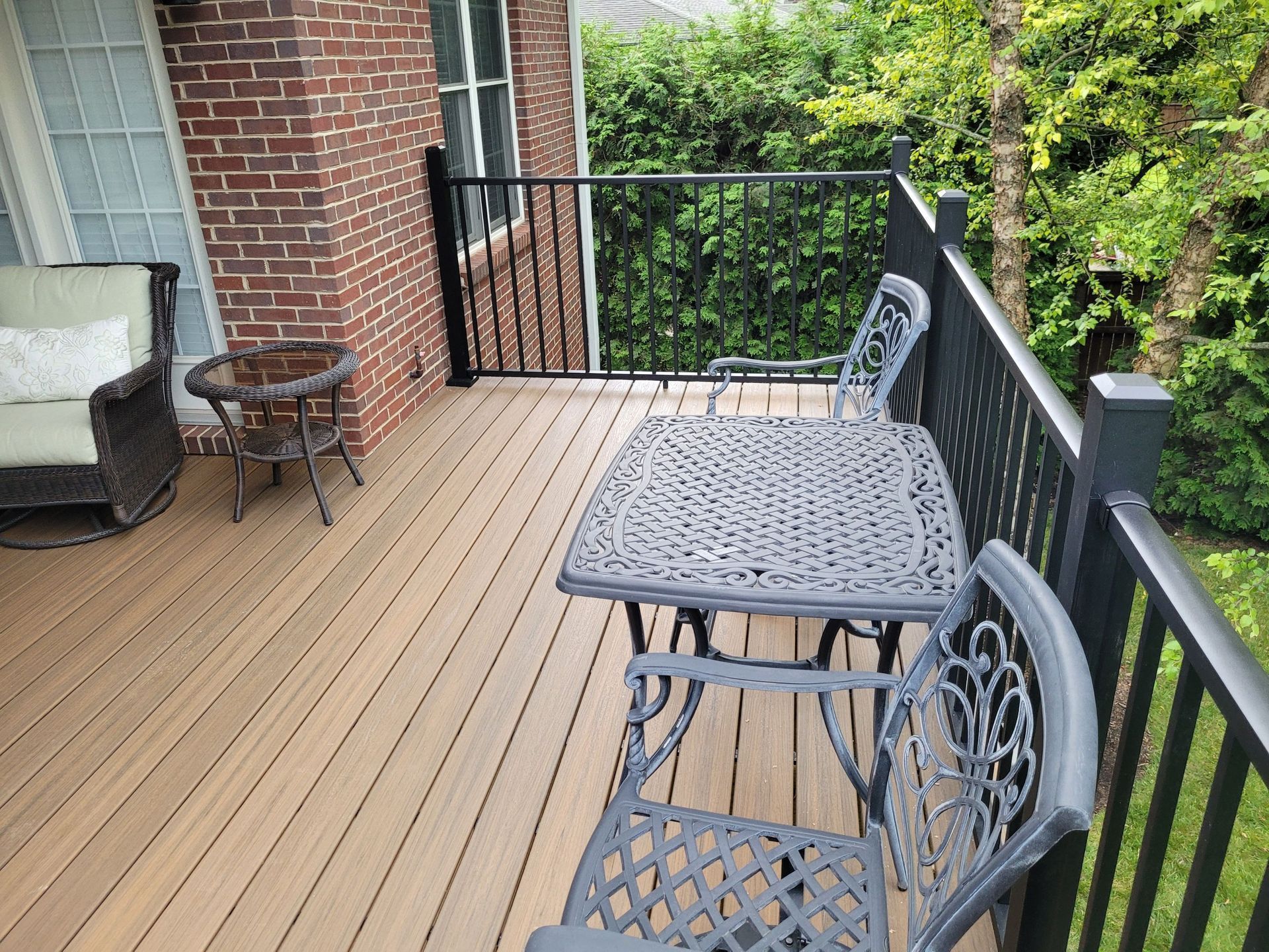 Brick patio deck with gray metal table and chairs beside a railing and green hedge