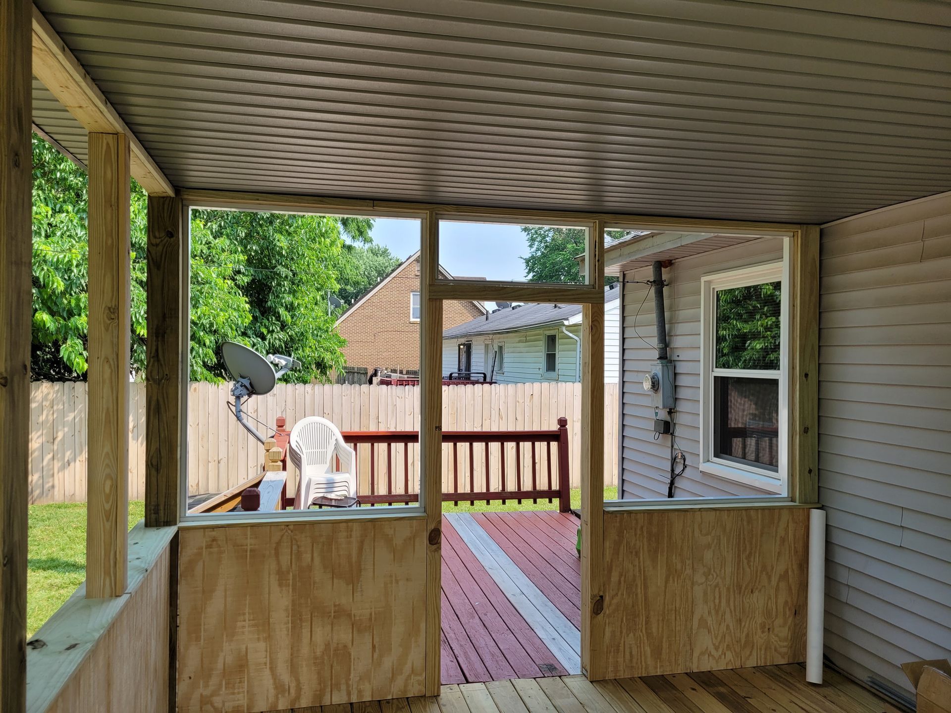 Covered backyard porch with plywood barriers, red deck, white chair, and a satellite dish view into a fenced yard