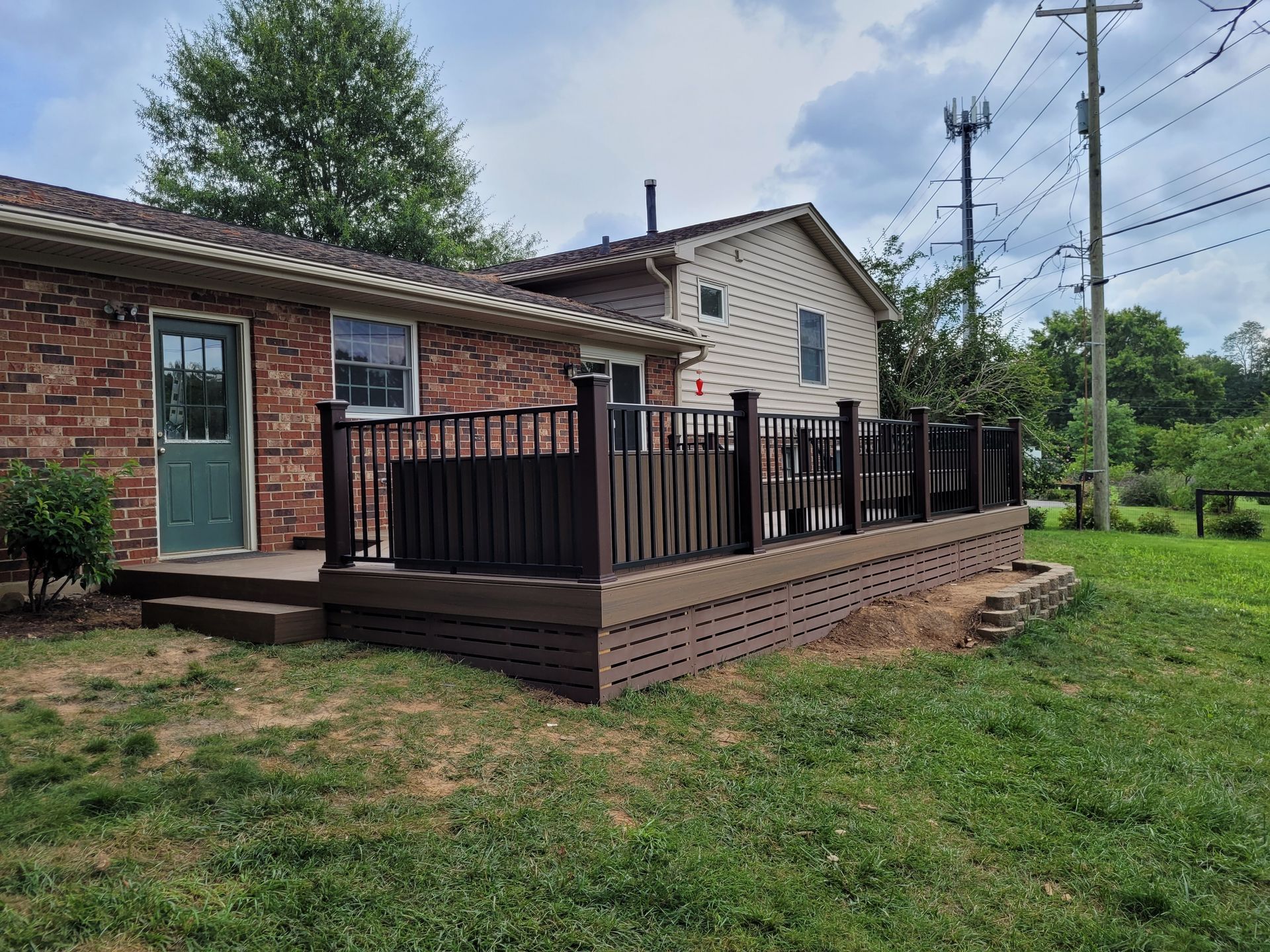 Small brick house with a green door and wooden deck railing beside a grassy yard under power lines.