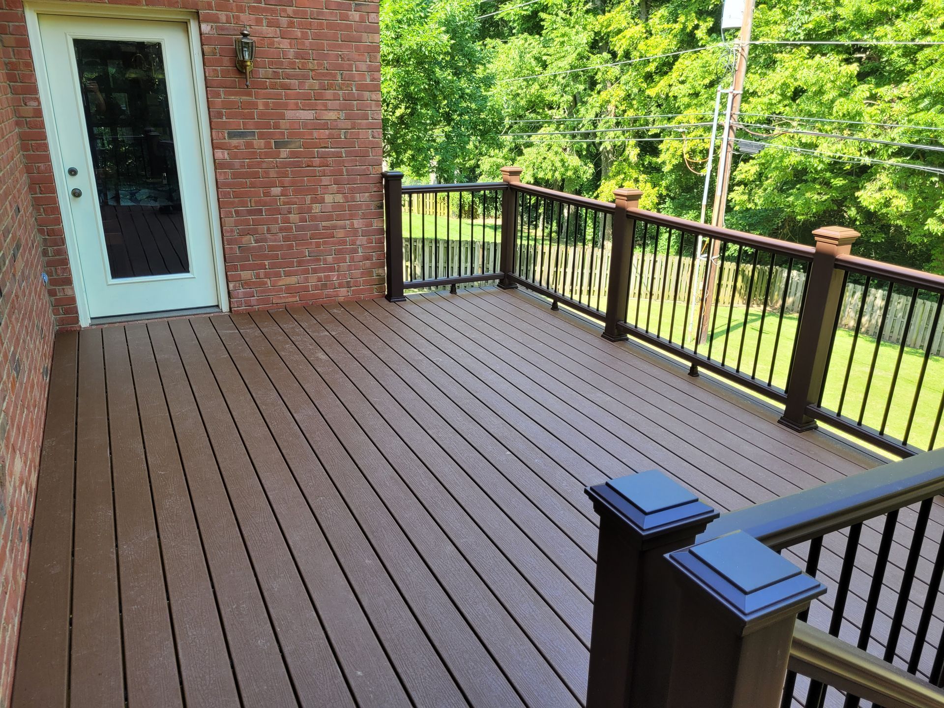 Brick patio deck with brown planks, black railings, and a glass door overlooking green trees