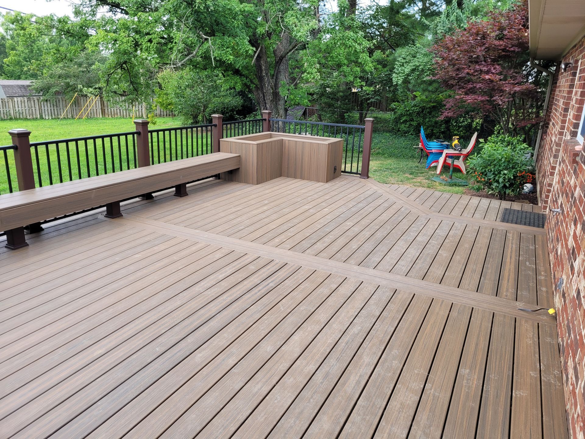 Wooden backyard deck with bench seating, a green lawn, and colorful chairs by the trees