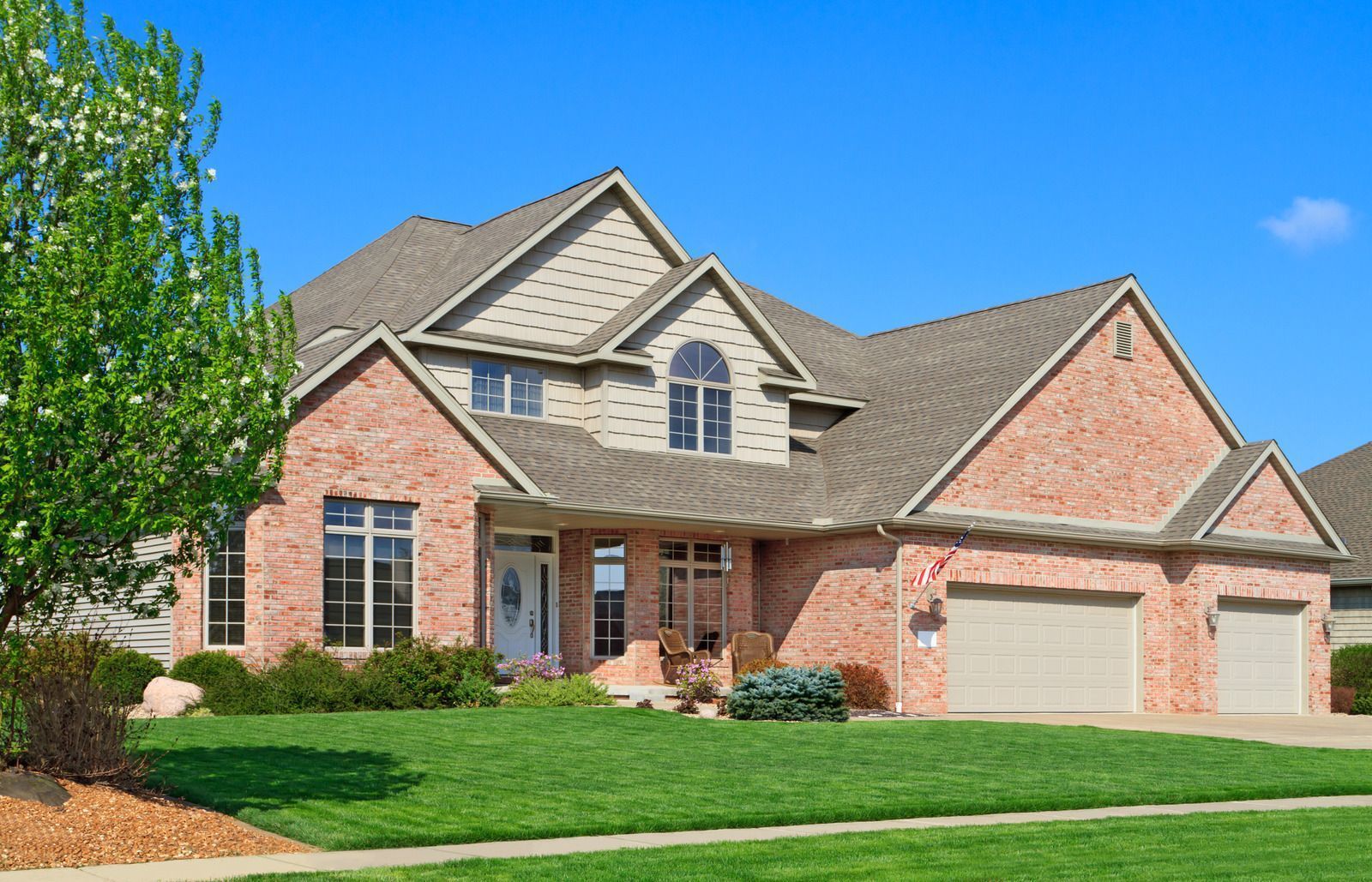 Suburban brick house with a two-car garage, gabled roof, and green lawn under a blue sky