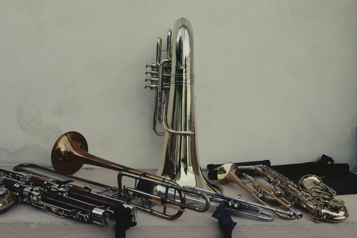 A collection of brass instruments, including a tuba and trombone, on a table against a white wall.