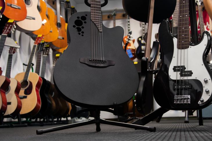 Black acoustic-electric guitar on a stand, in a store with many other guitars in the background.