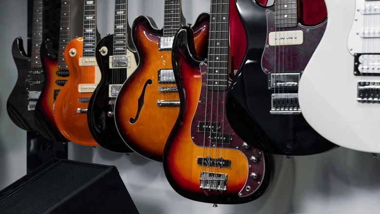 A row of six electric guitars, including a bass, in various colors, hanging on a wall.
