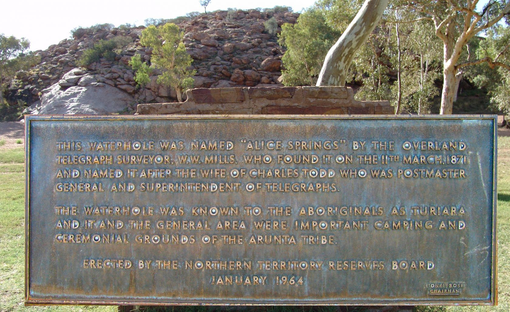 Bronze plaque with text, set outdoors, with a rocky hillside in the background.