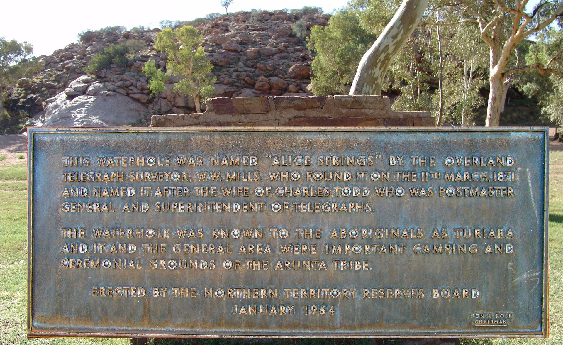 Bronze plaque with inscription in front of a rocky landscape, green grass, and trees.