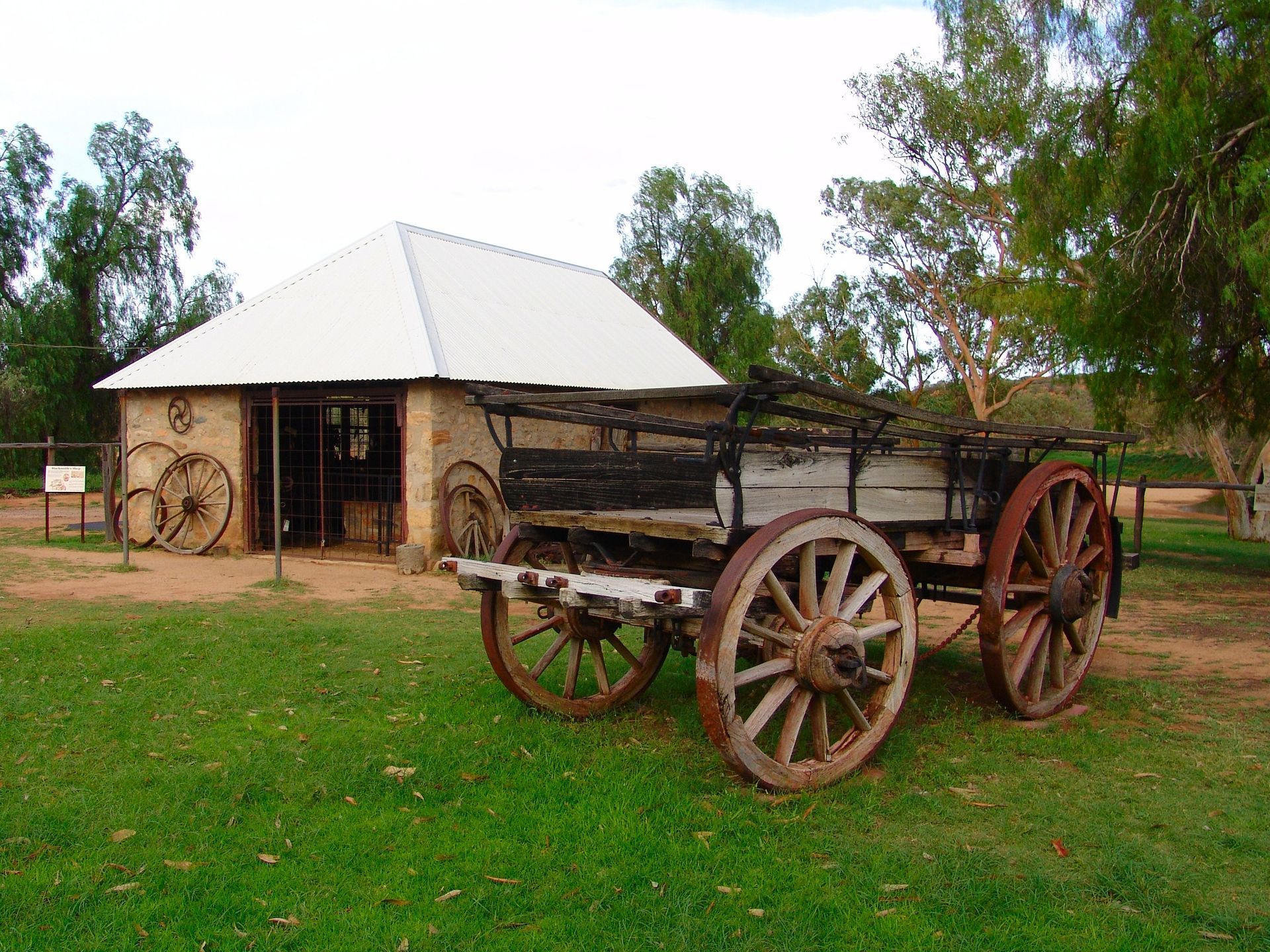 Old wooden wagon in front of a stone building with metal roof; green grass, trees in background.