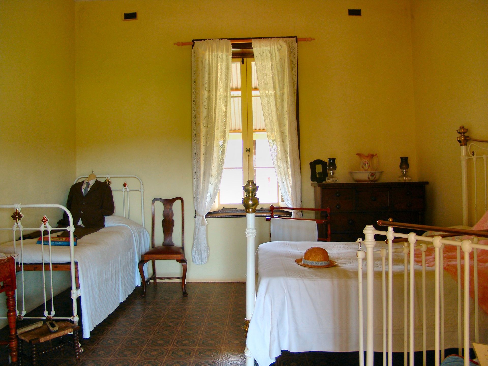 Bedroom with two antique white beds, wooden chair, and window with lace curtains.