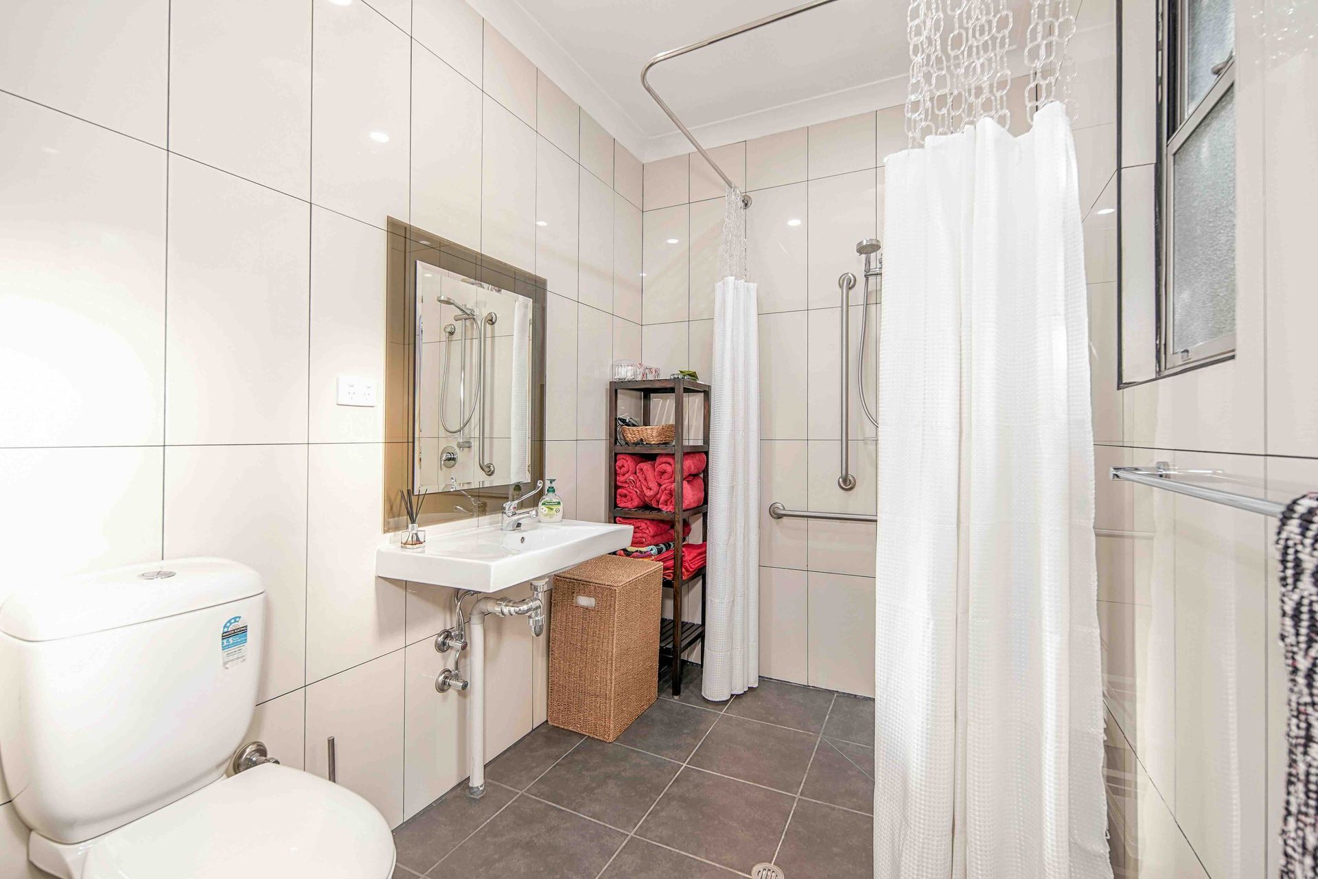 Bathroom with white tile walls, toilet, sink, shower with curtain, and storage shelf.