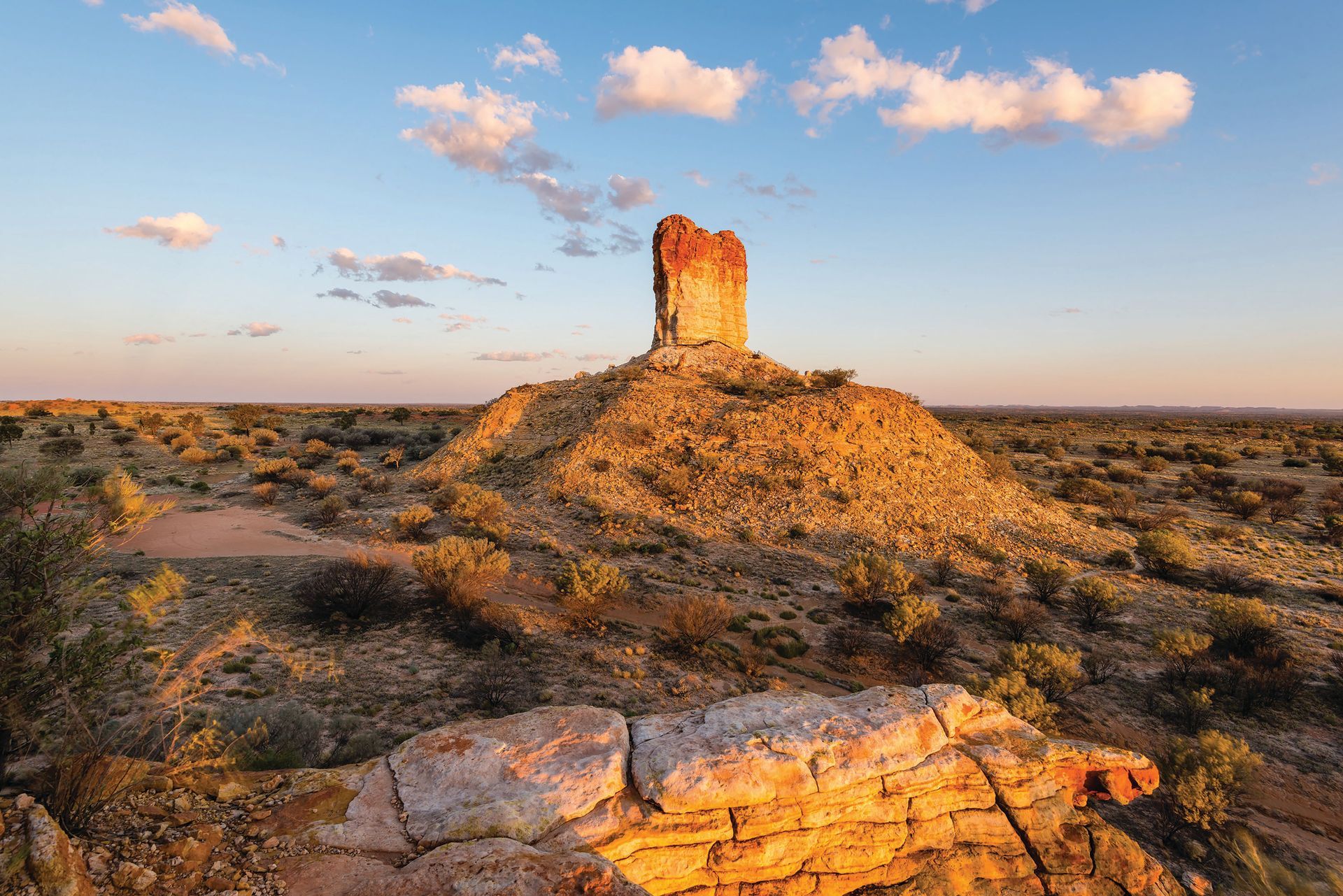 A tall, reddish rock formation rises from a desert landscape under a blue and cloudy sky.
