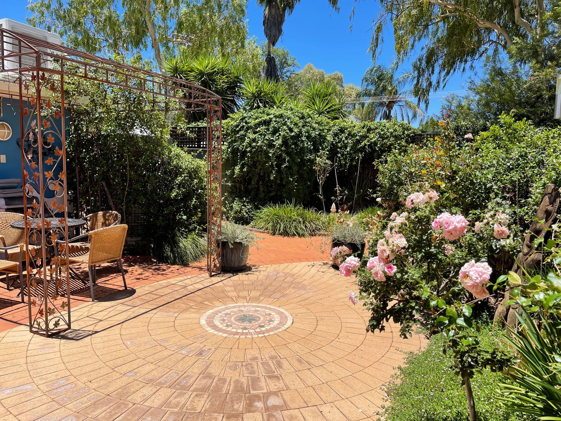 Brick patio in a lush garden with rose bushes, shaded seating area under a metal arbor, and blue sky.