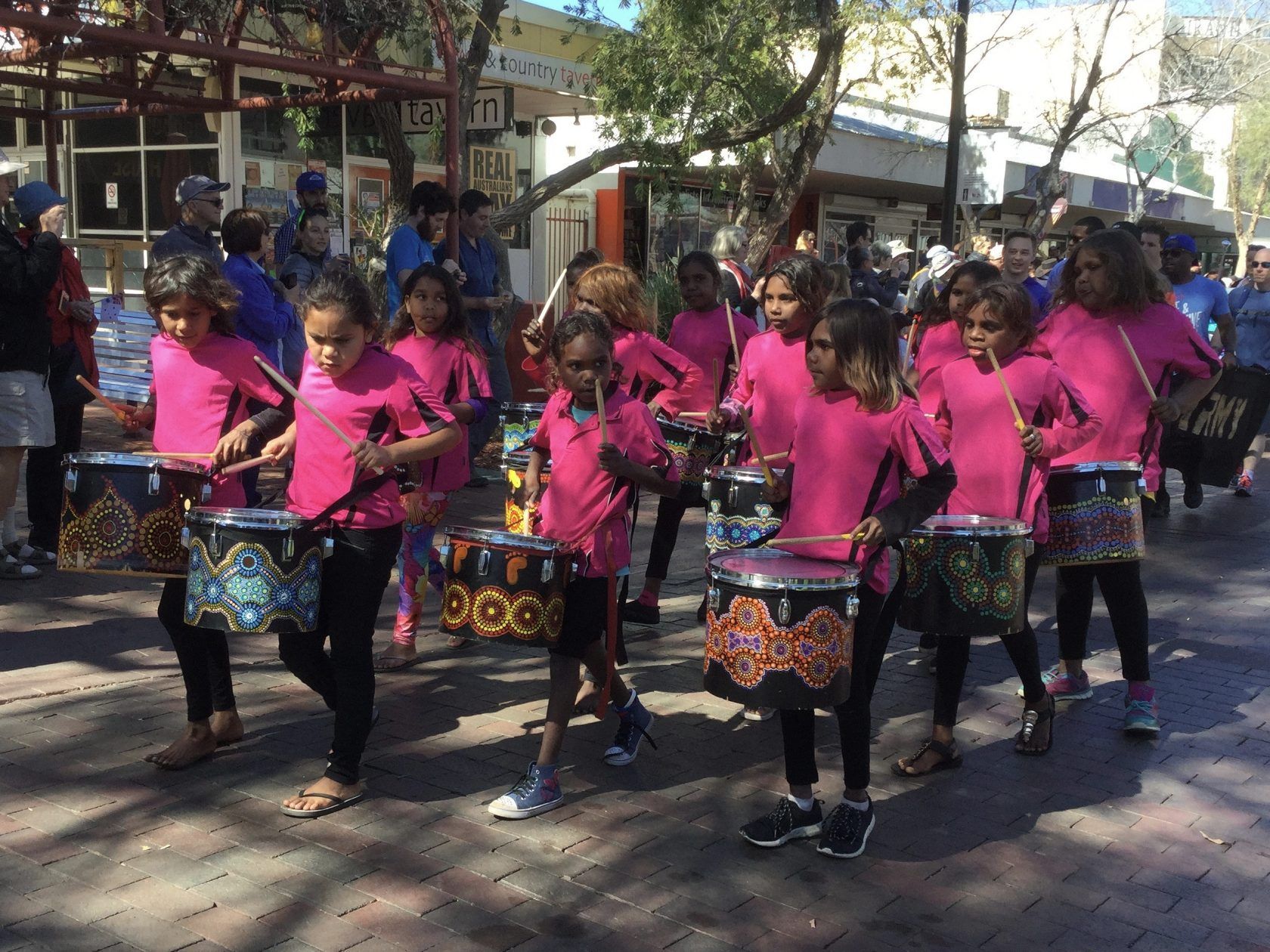 Group of children in pink shirts playing drums in a street parade.