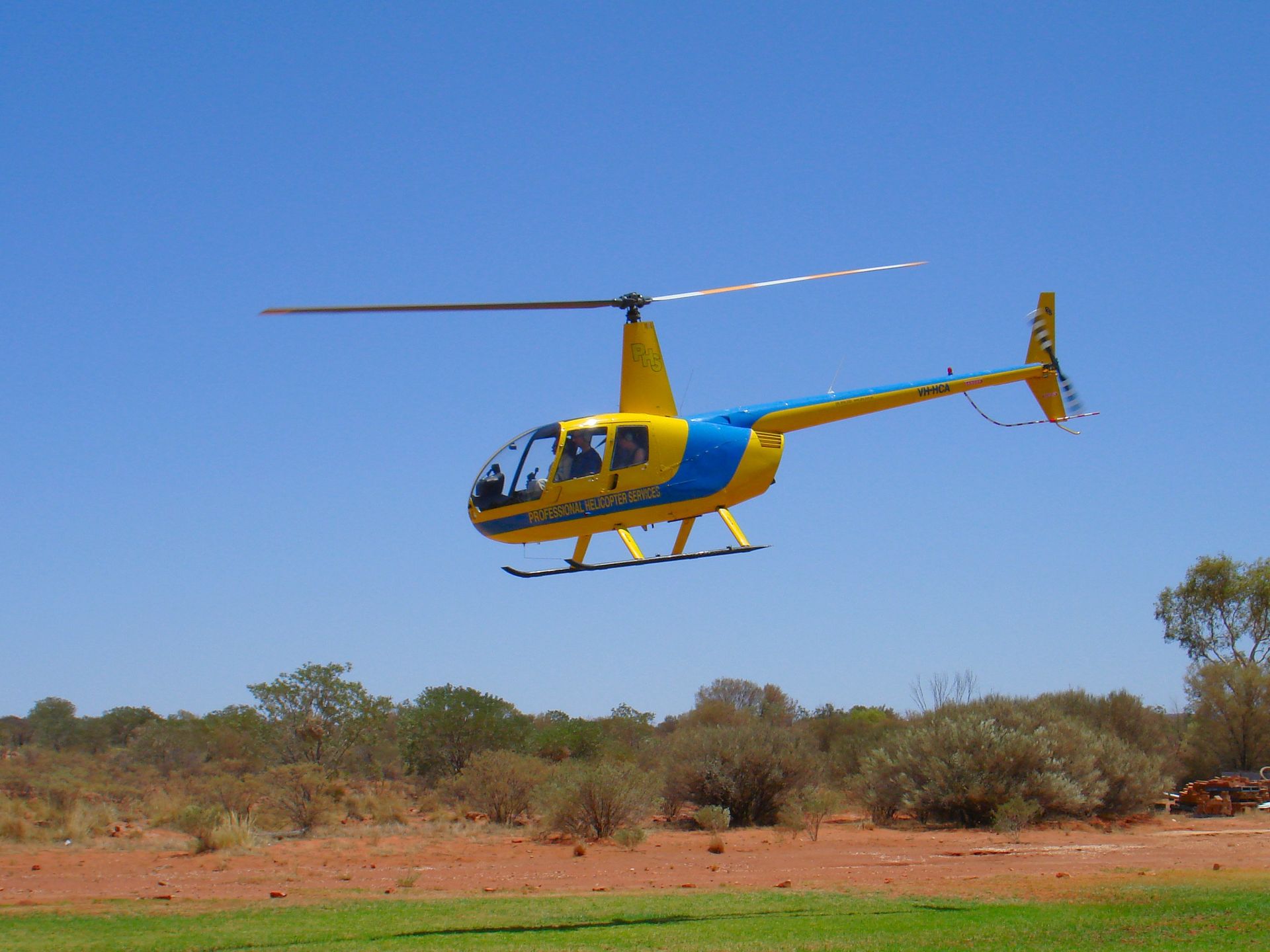 Yellow and blue helicopter taking off over red dirt and shrubs, with a blue sky.