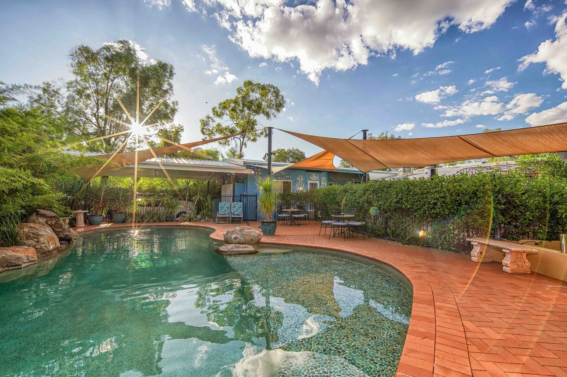 Poolside area with pool, brick patio, sunshades, and hedges under a sunny sky.