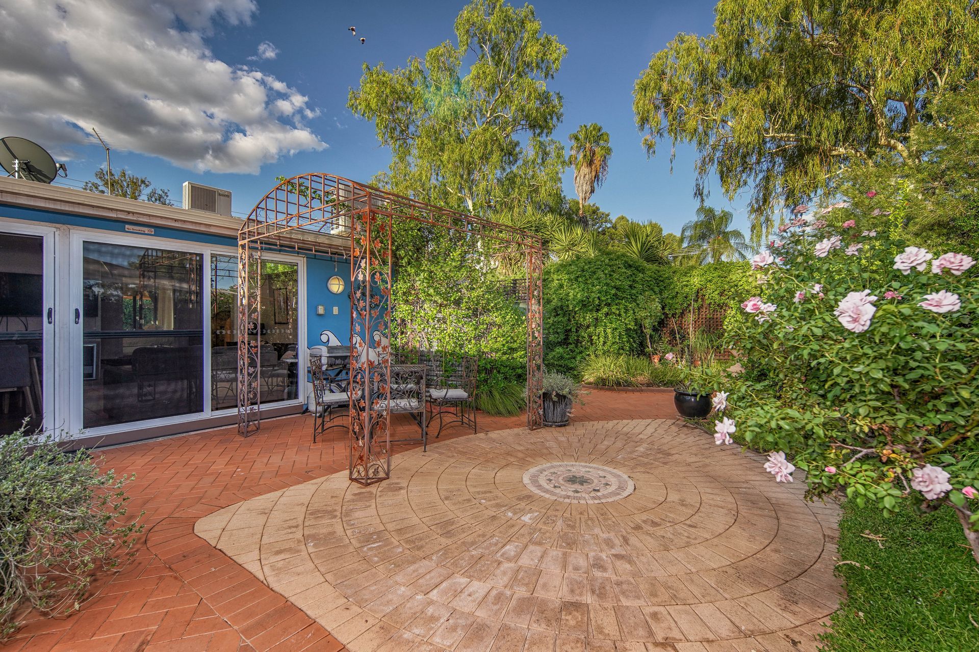 Brick patio with metal arbor, chairs, and lush garden. Building with glass doors in background.