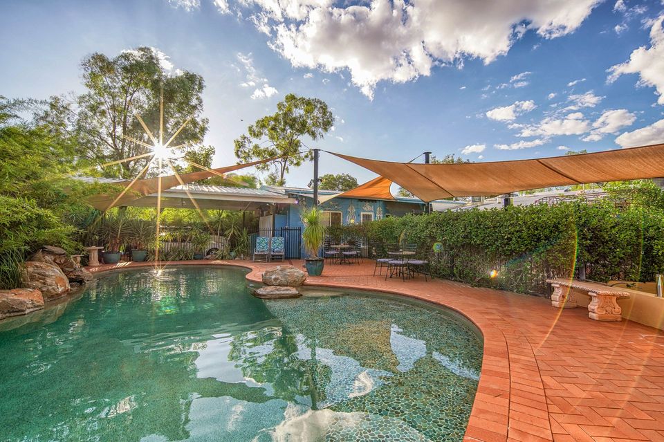 Poolside scene with a curved pool, red brick path, shade sails, and lush greenery under a sunny sky.