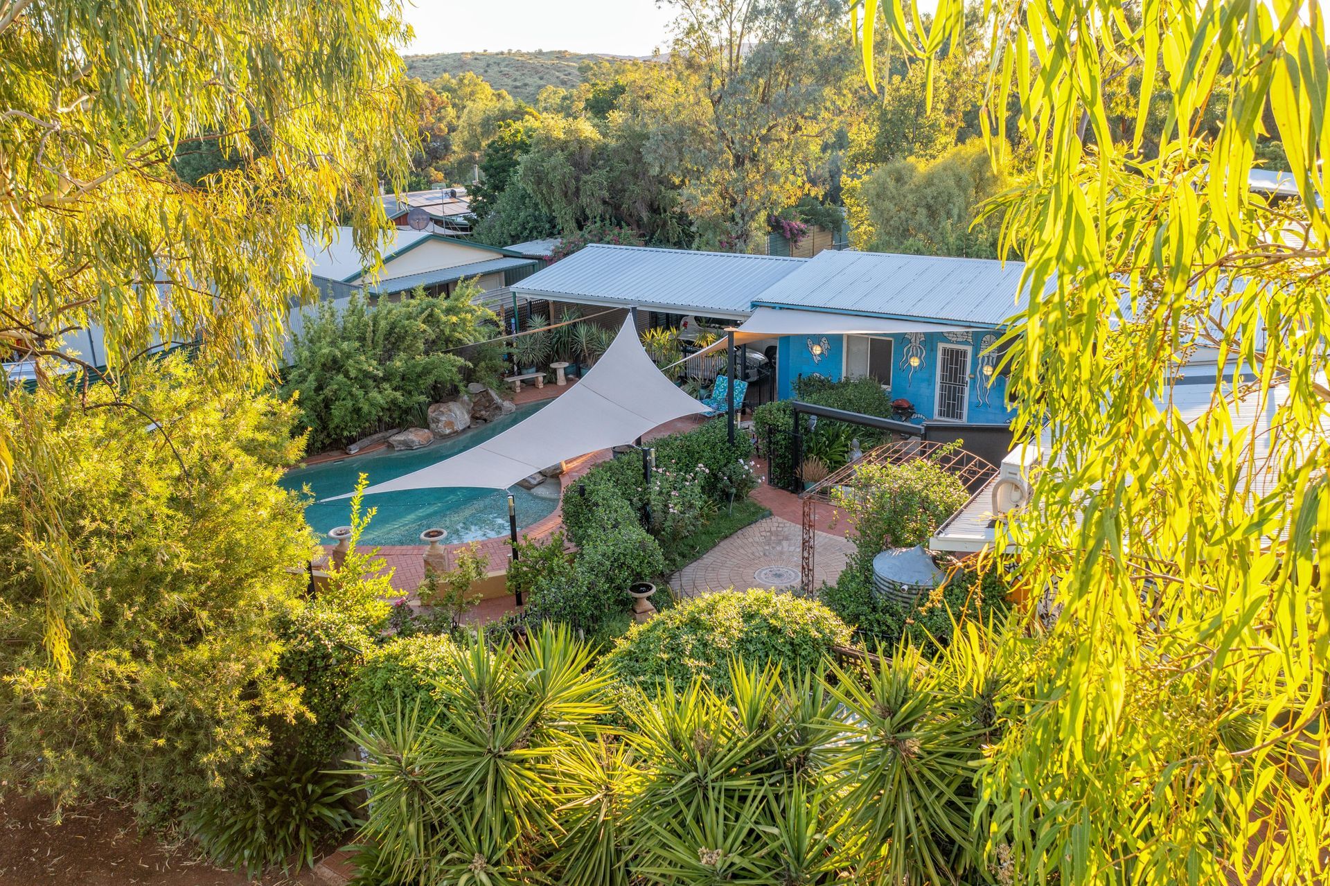 A blue house with a pool and shade sail, surrounded by lush green trees in a sunny setting.