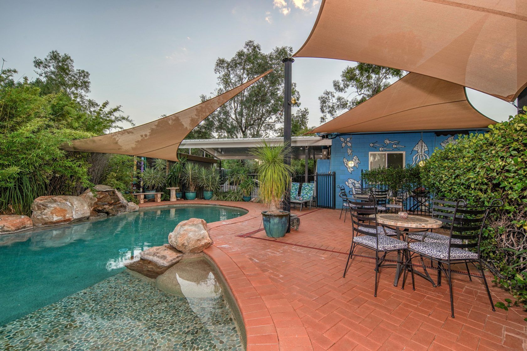 Backyard pool and patio with shade sails, blue building, and greenery.